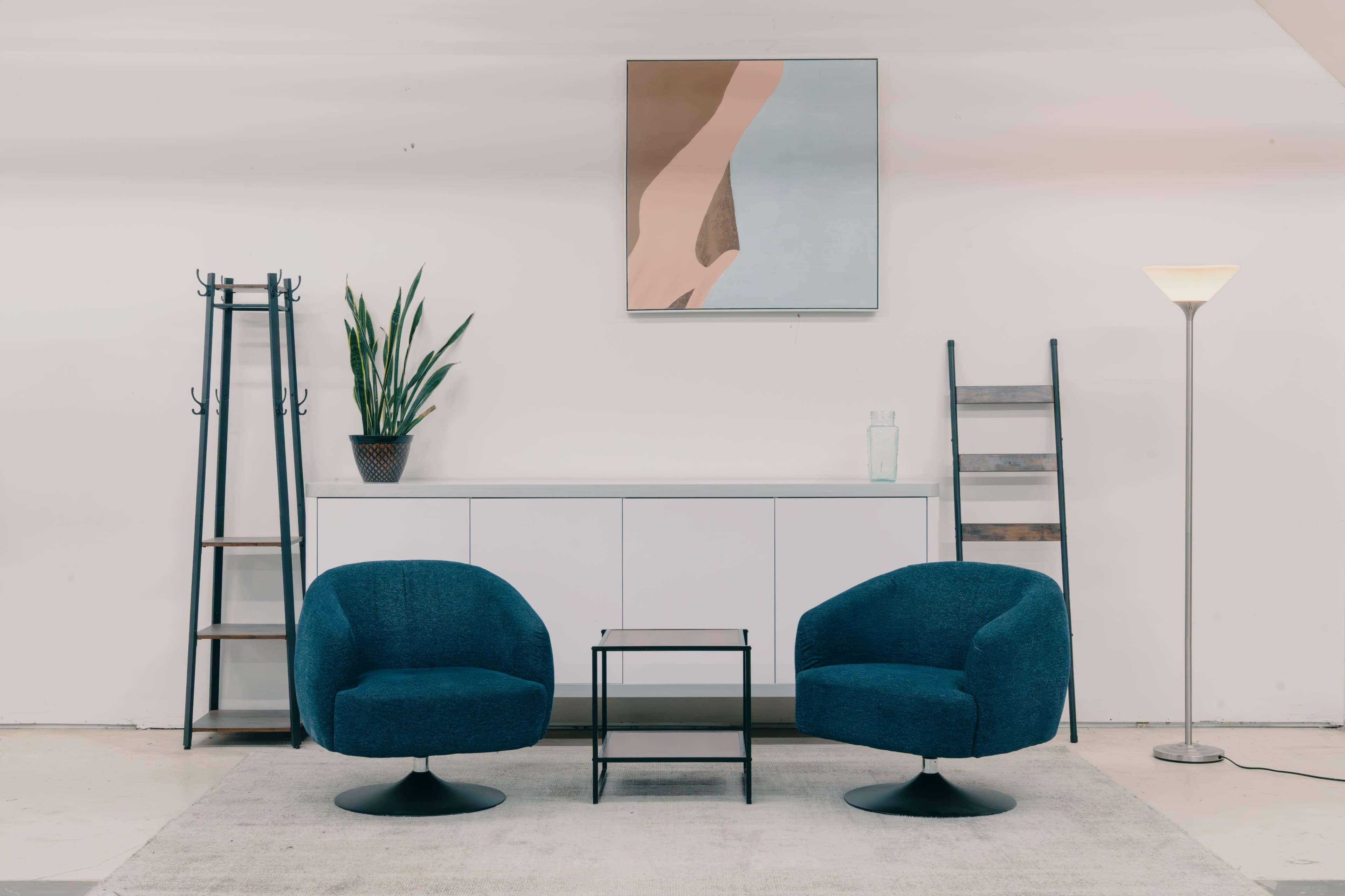 The image shows a minimalist interior featuring two blue chairs, a small black table, a plant, a white cabinet, a painting on the wall, and a ladder-style shelf.
