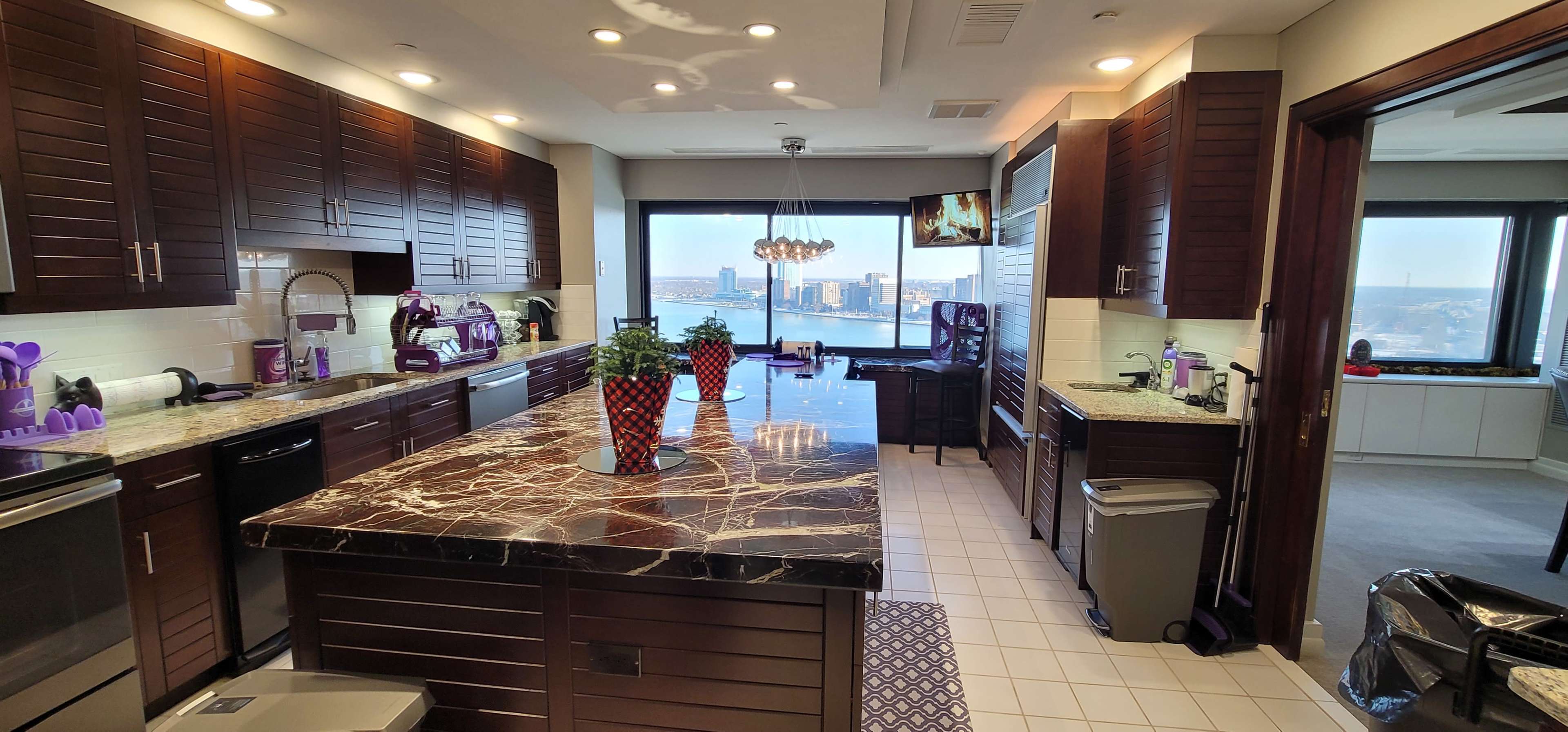 The image shows a modern kitchen with dark wooden cabinetry, a large marble island, and a view of the city through a window.