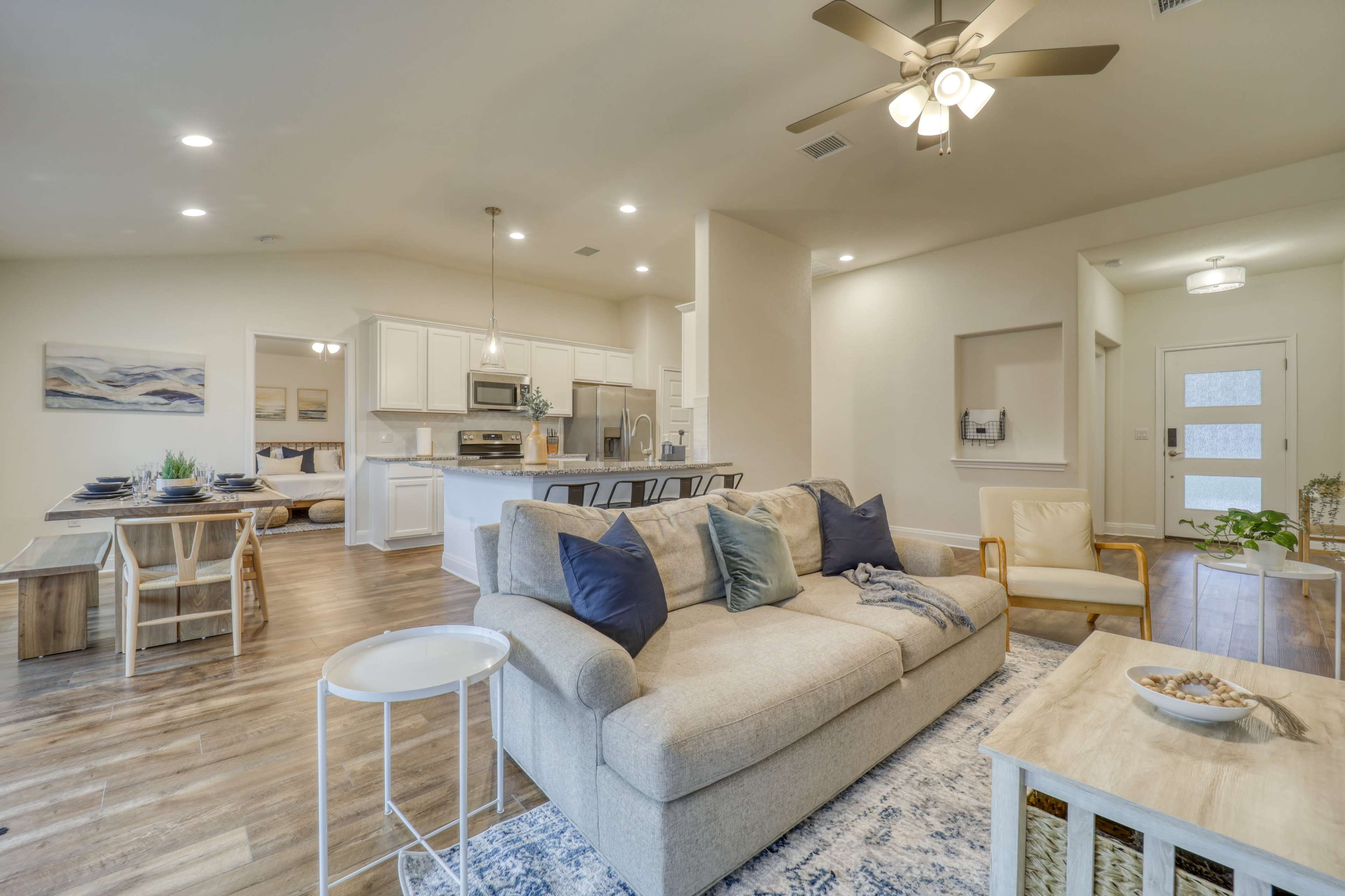 The image shows a bright and open living area with a beige sofa, a wooden dining table, and modern kitchen appliances.