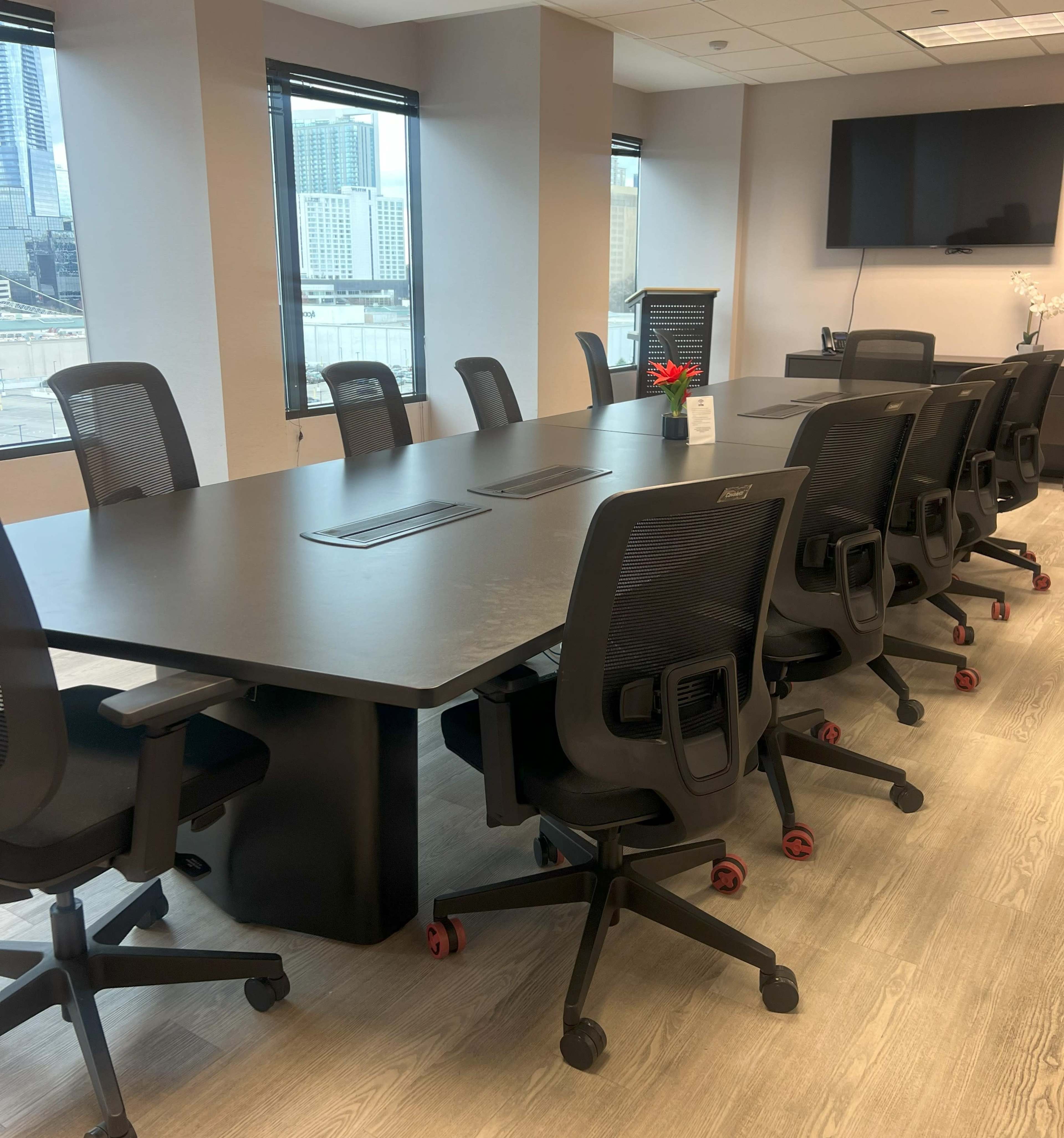 A modern conference room features a large rectangular table surrounded by ergonomic chairs and large windows offering a view of the city.
