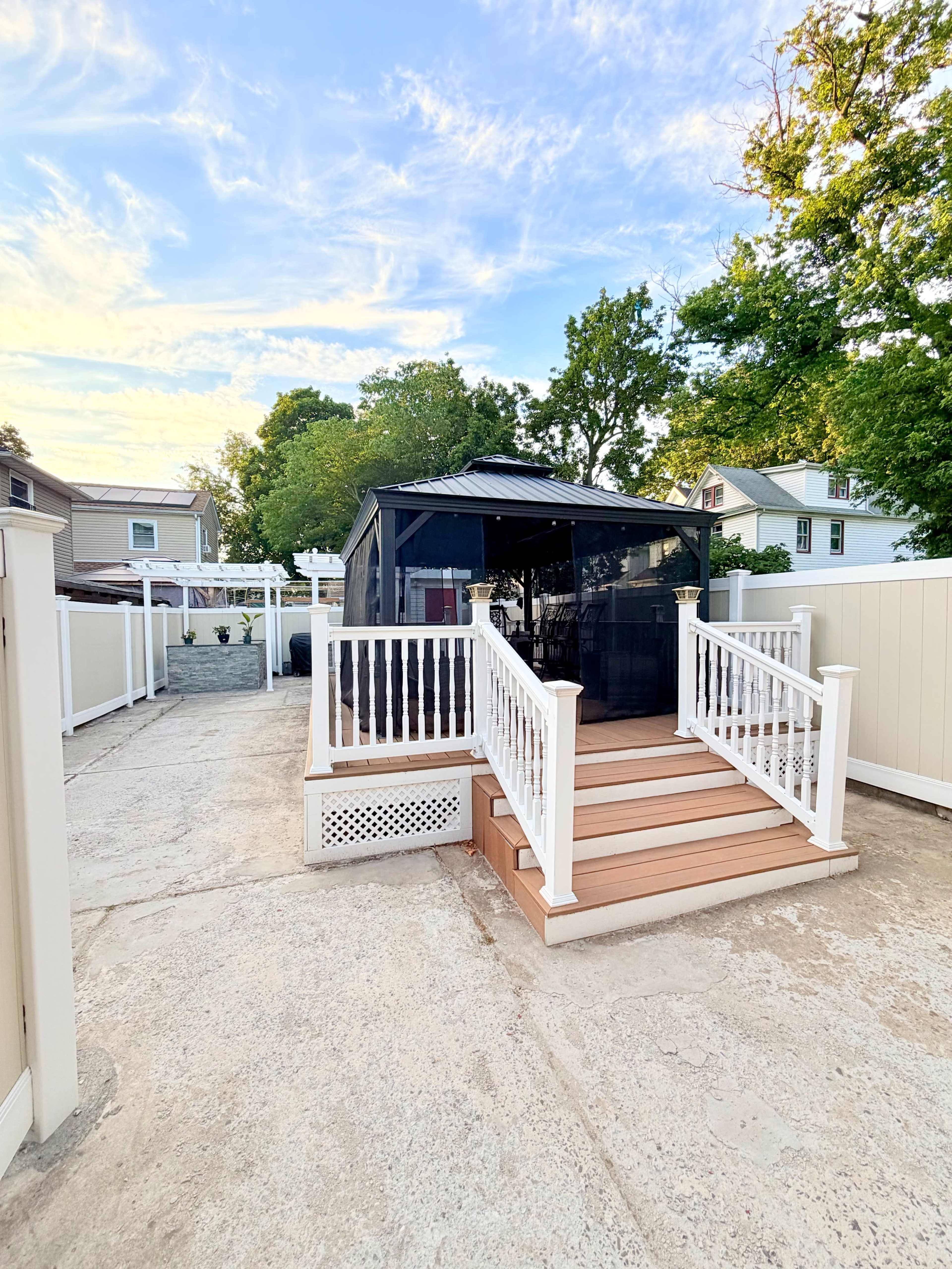 A gazebo with a black canopy is situated on a wooden deck, surrounded by a concrete patio and white fences under a clear sky.