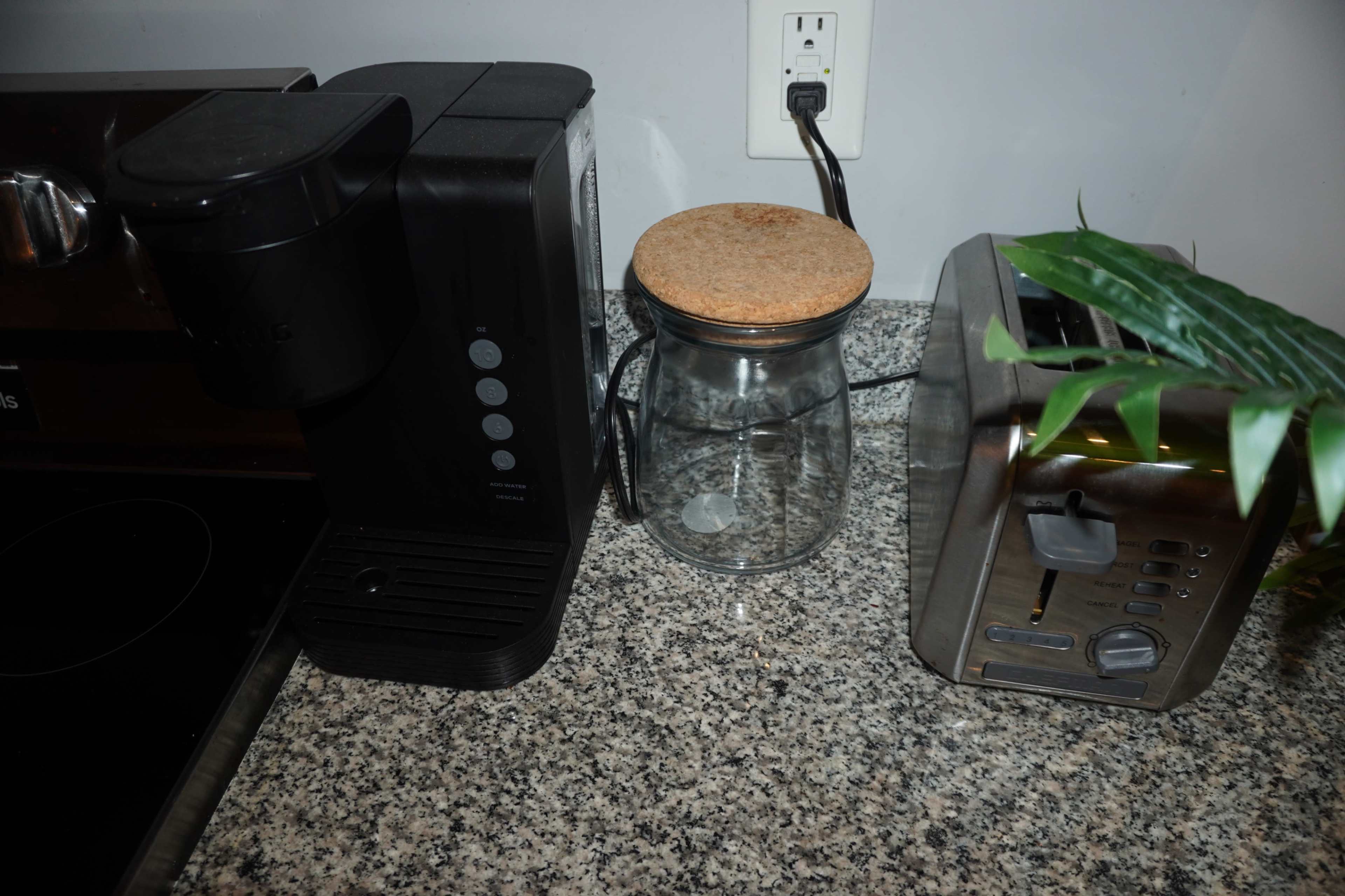 The image shows a coffee maker and a toaster next to a glass jar with a cork lid, all placed on a granite countertop.