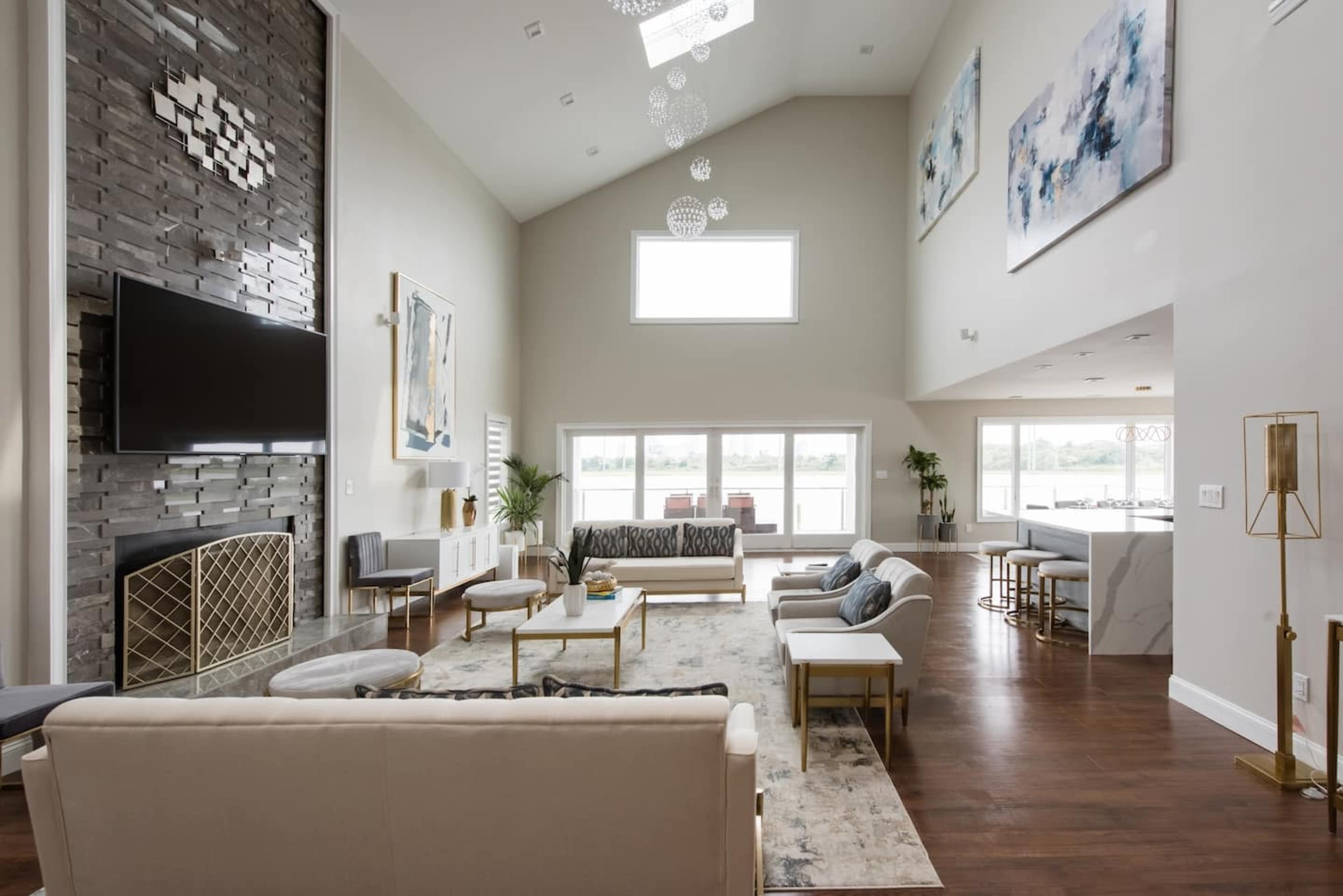 The scene features a spacious living room with high ceilings, large windows, and modern furnishings arranged around a fireplace and a central area rug.