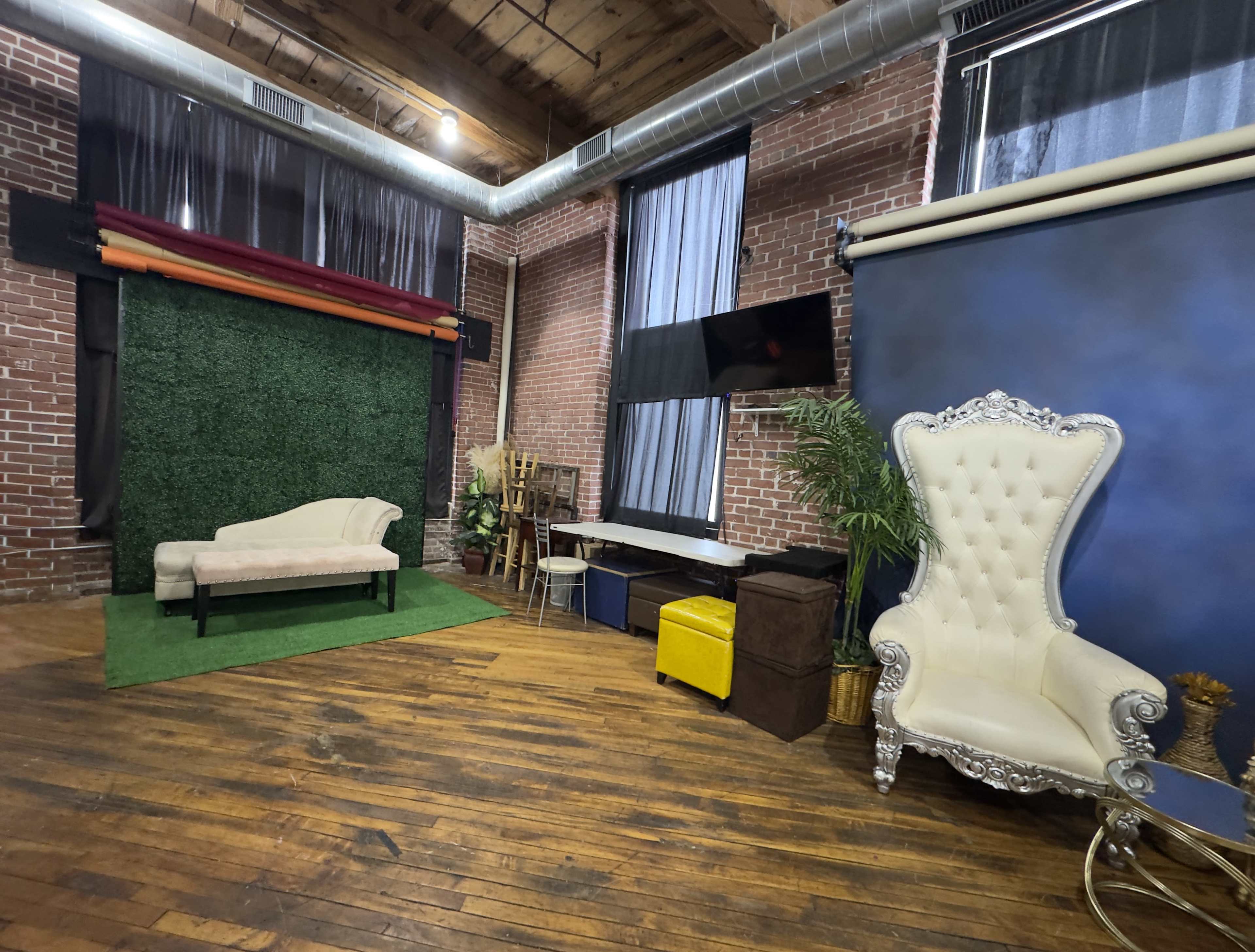 The image shows a spacious studio with a wooden floor, featuring a green backdrop area, a light-colored chaise lounge, a white ornate chair, and various props arranged around the room.