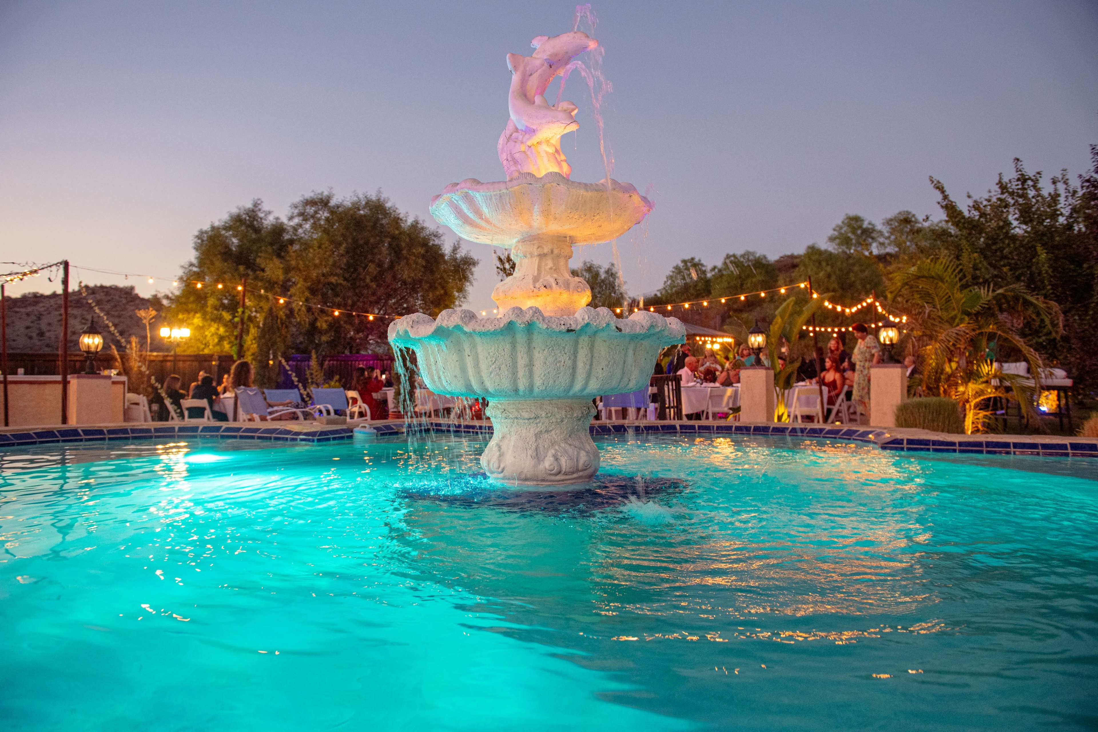 A decorative fountain with a statue of a cherub is illuminated at dusk, surrounded by a pool of blue water and outdoor seating.