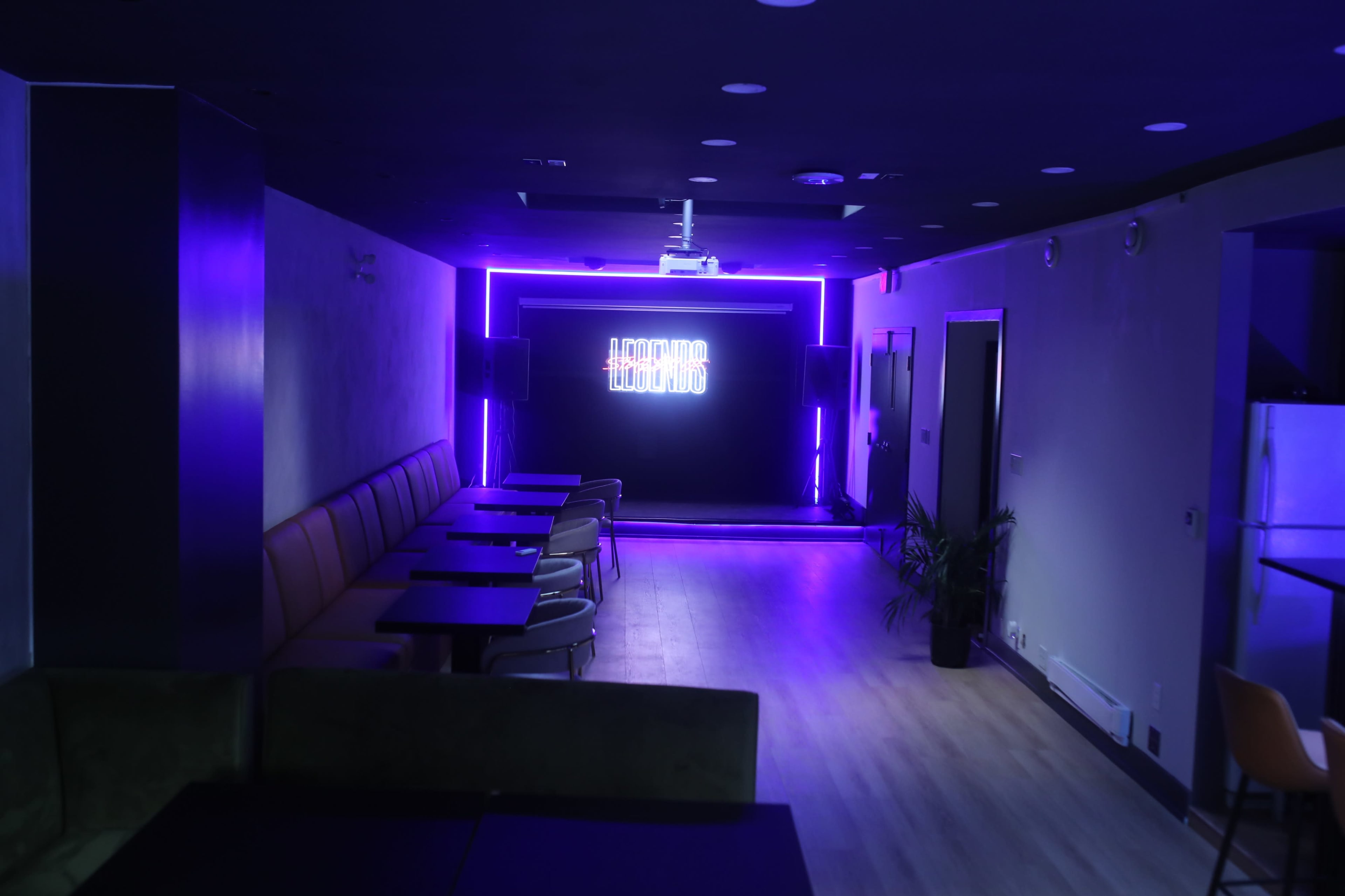 The image shows a dimly lit lounge area with purple neon lighting and rows of tables arranged in front of a screen.