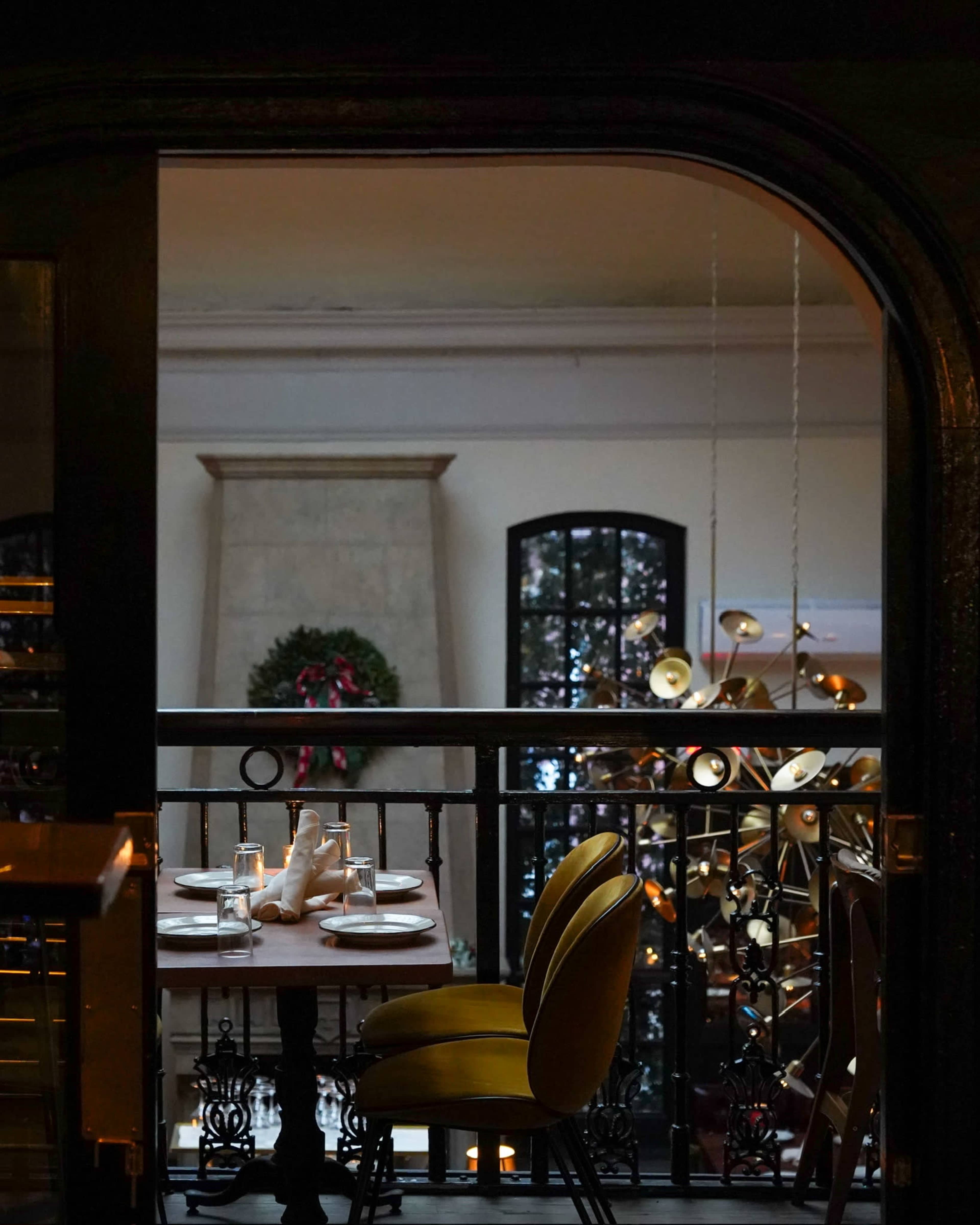 A dining table set with plates and napkins, positioned next to a wall with an archway, overlooking a decorative space with a chandelier and a holiday wreath.