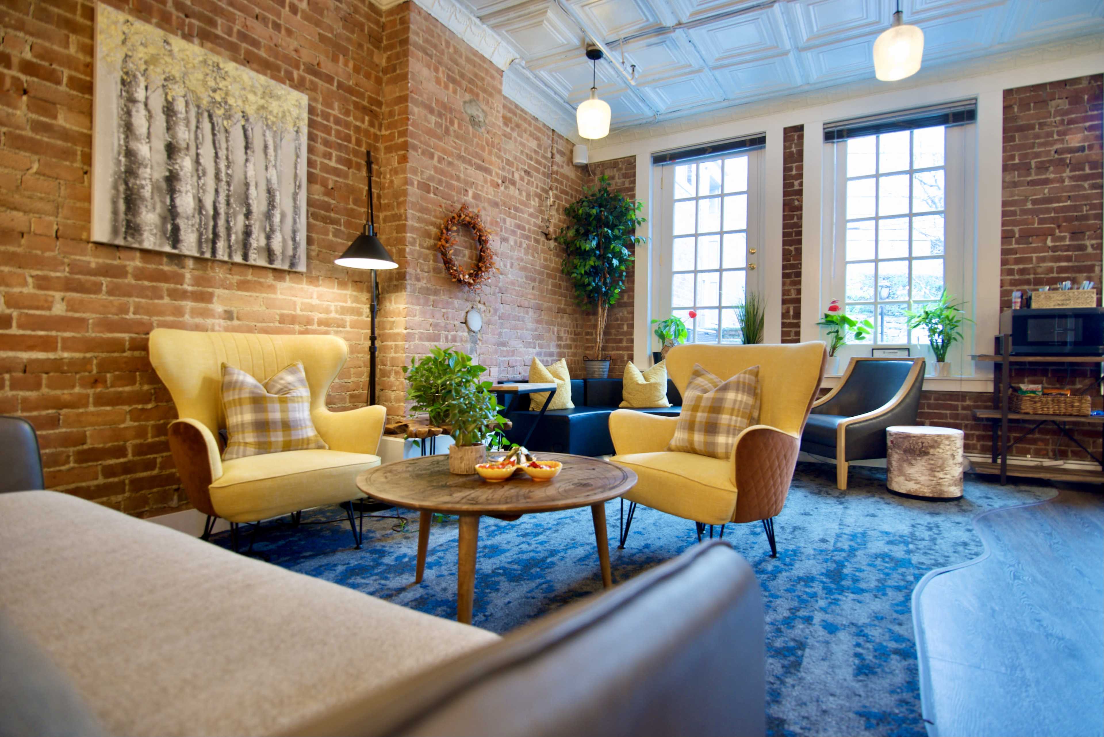 A cozy sitting area with yellow chairs, a wooden table, and large windows surrounded by exposed brick walls and plants.