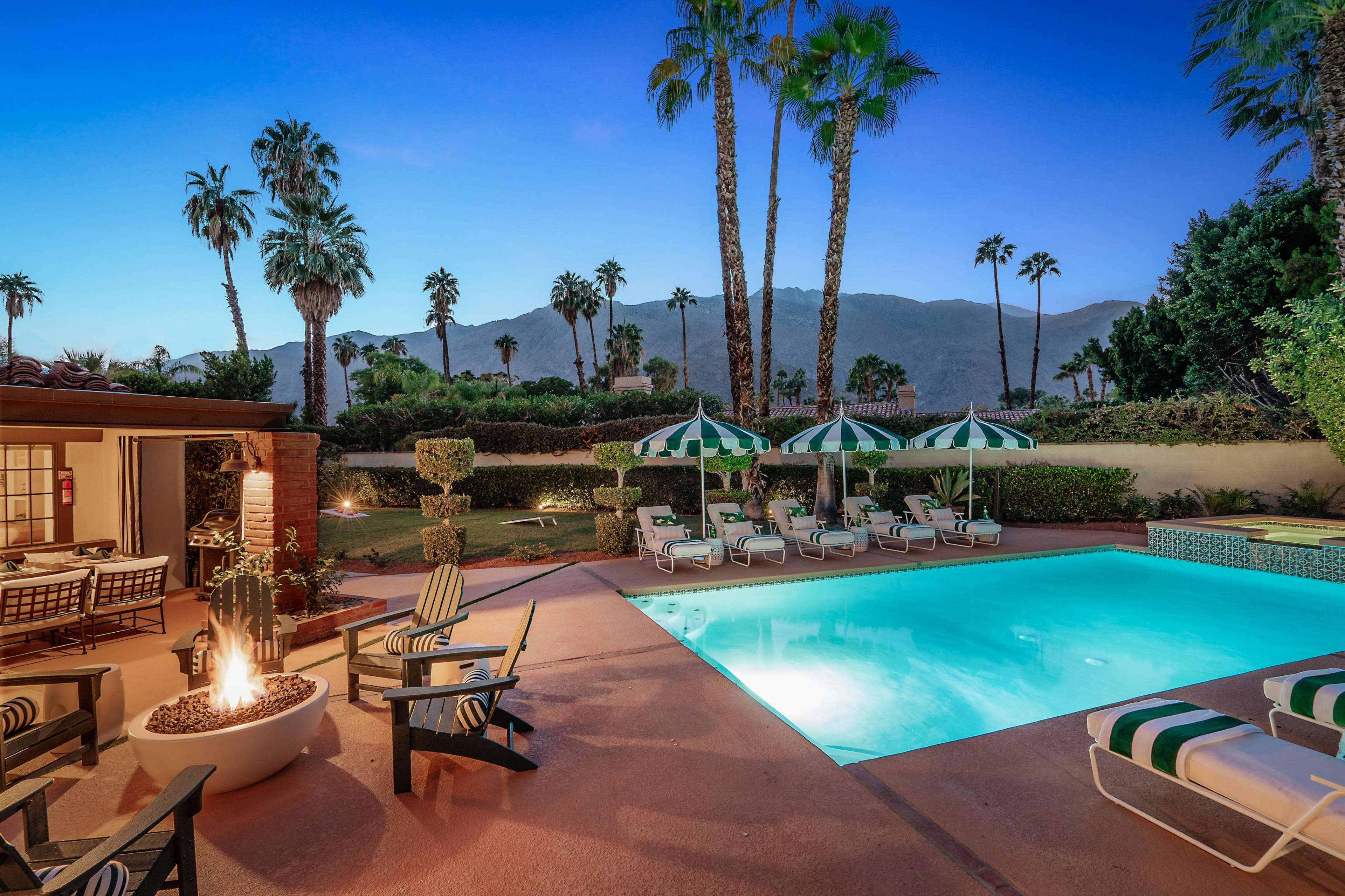 The image shows a pool area surrounded by palm trees, with lounge chairs and striped umbrellas, set against a backdrop of mountains at dusk.