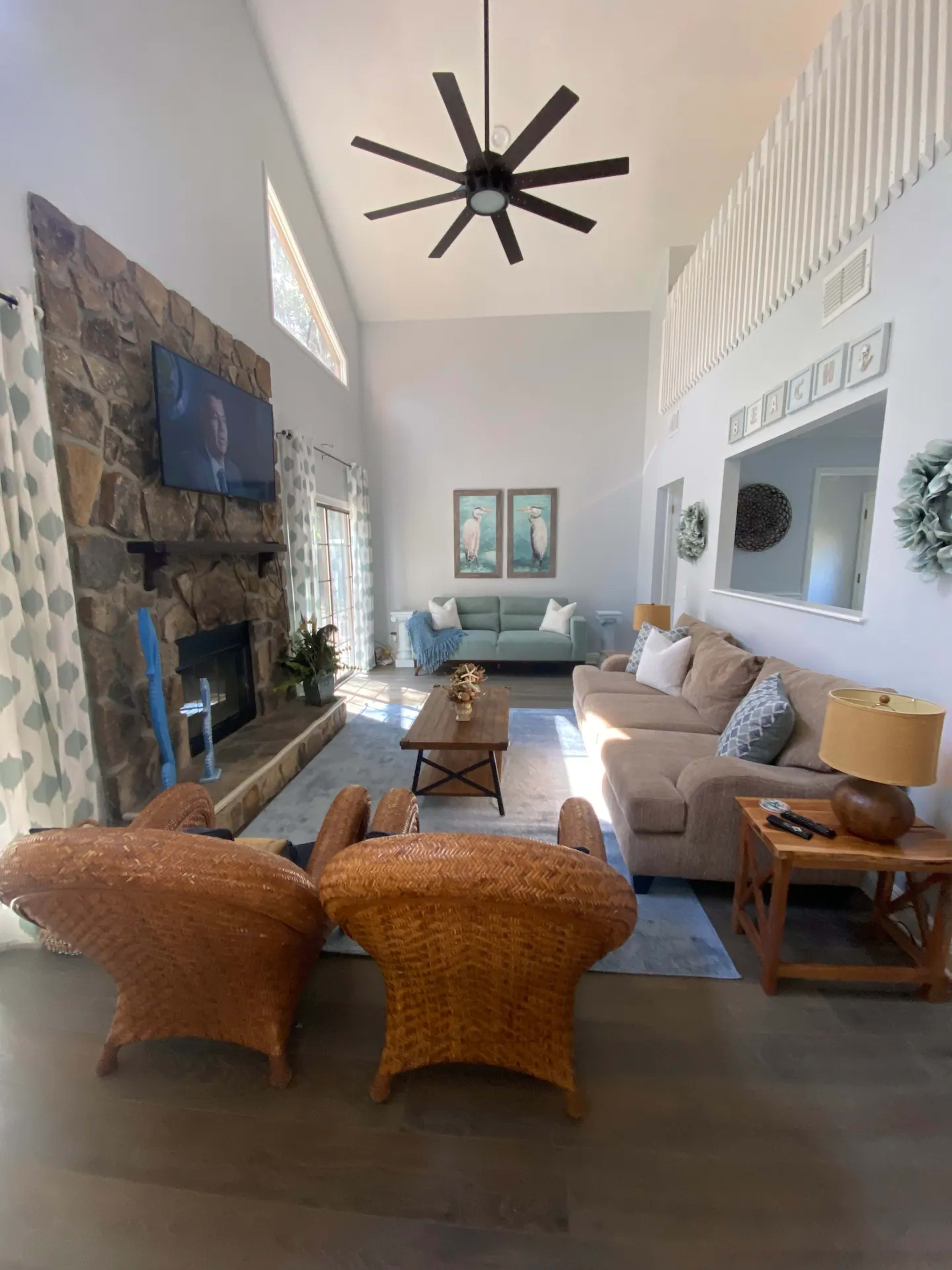 The image shows a spacious living room featuring a stone fireplace, a large sofa, two woven armchairs, and a television mounted on the wall.