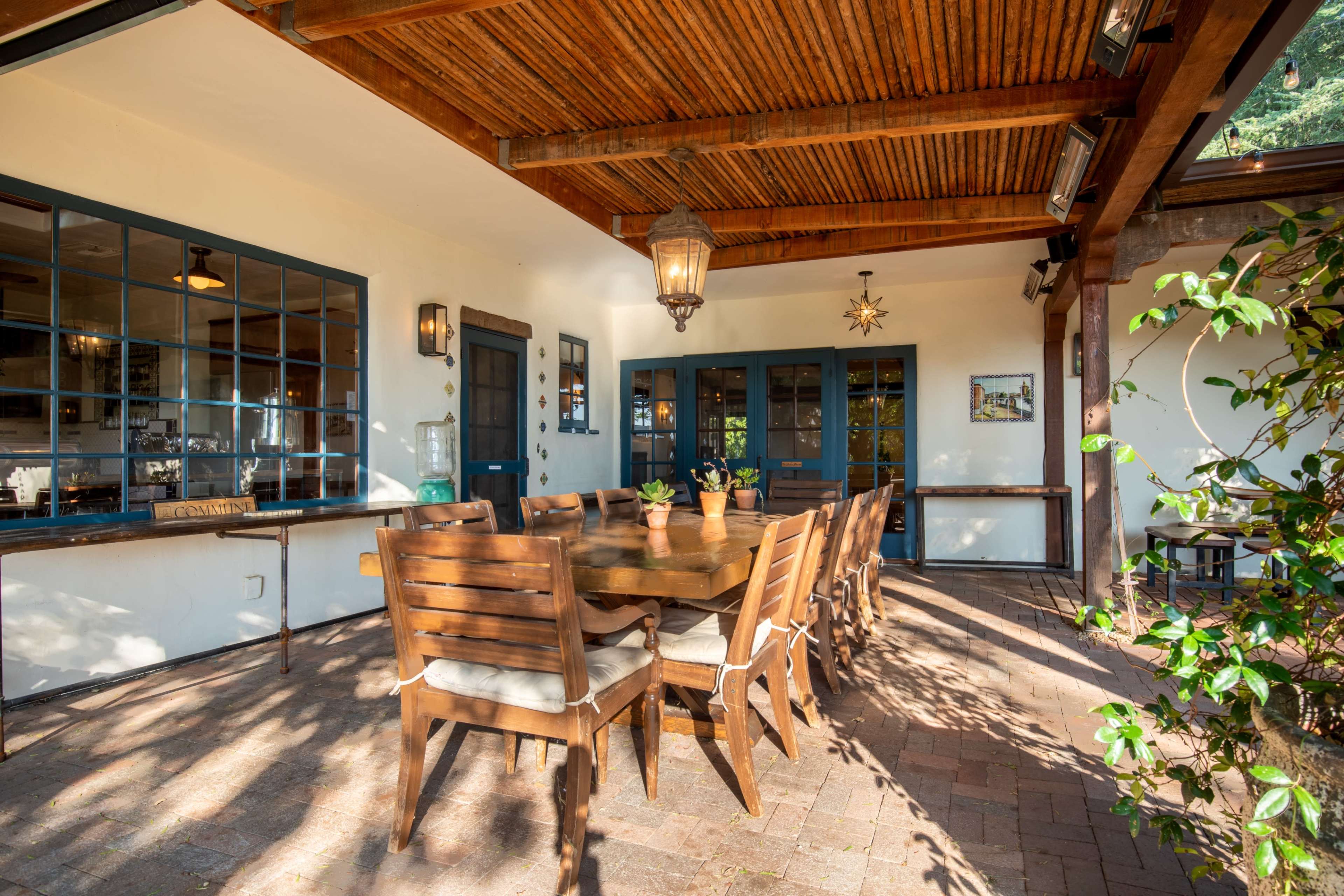 A dining area with a large wooden table and chairs is set under a covered patio, surrounded by plants and with light streaming through.