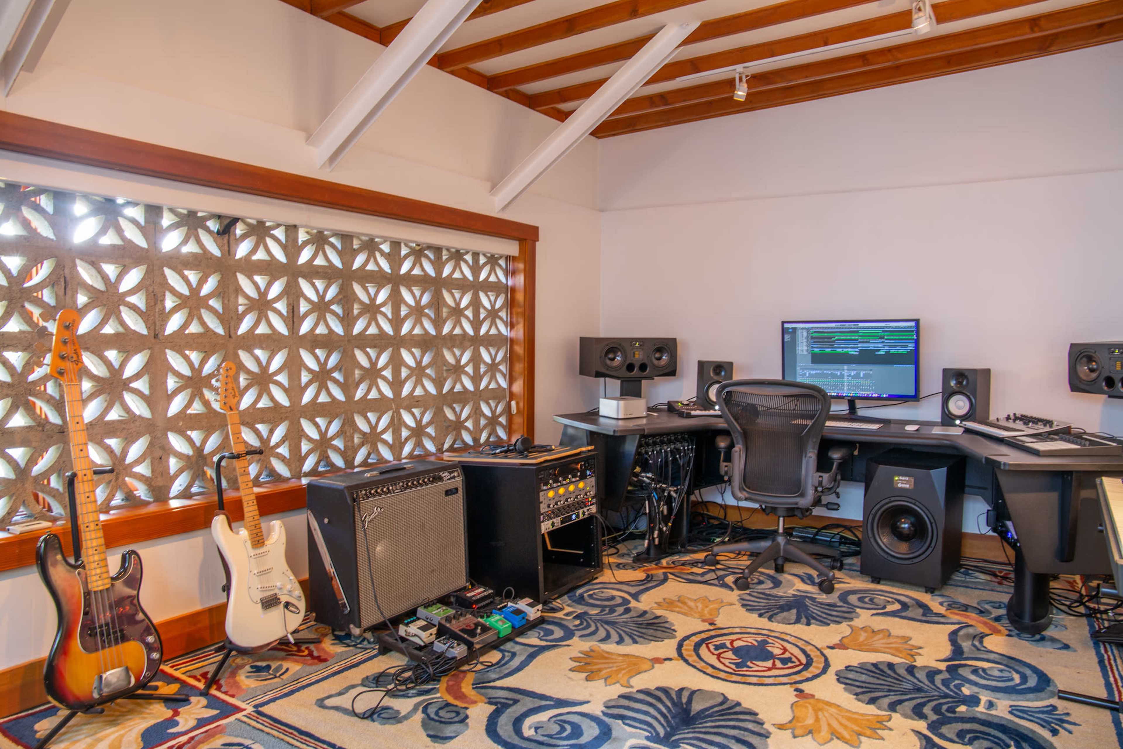A music production studio with guitars, amplifiers, and a computer setup, featuring a decorative window.