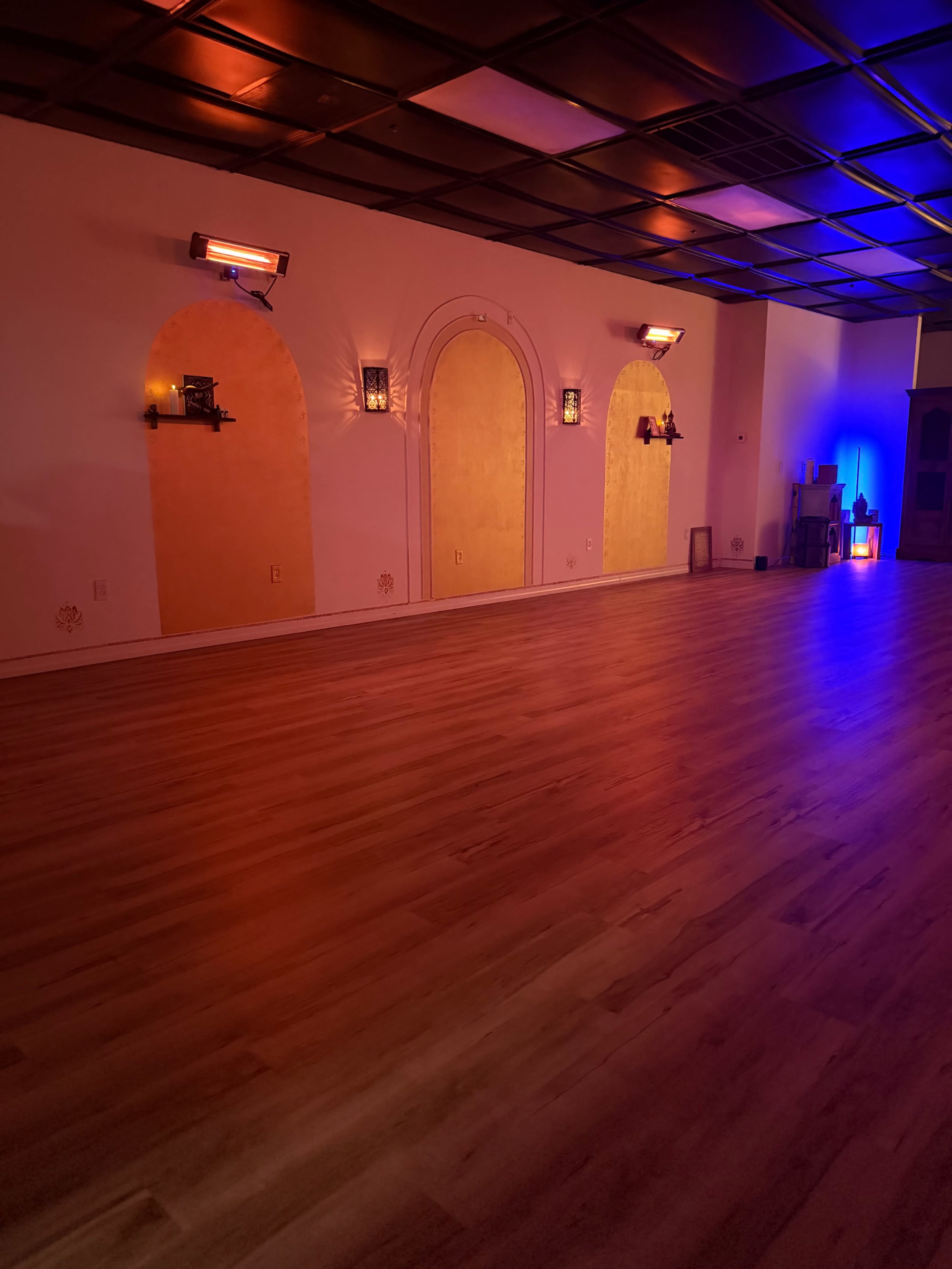 The image shows an empty yoga studio with wooden flooring, three arched niches on the wall, and ambient lighting in various colors.