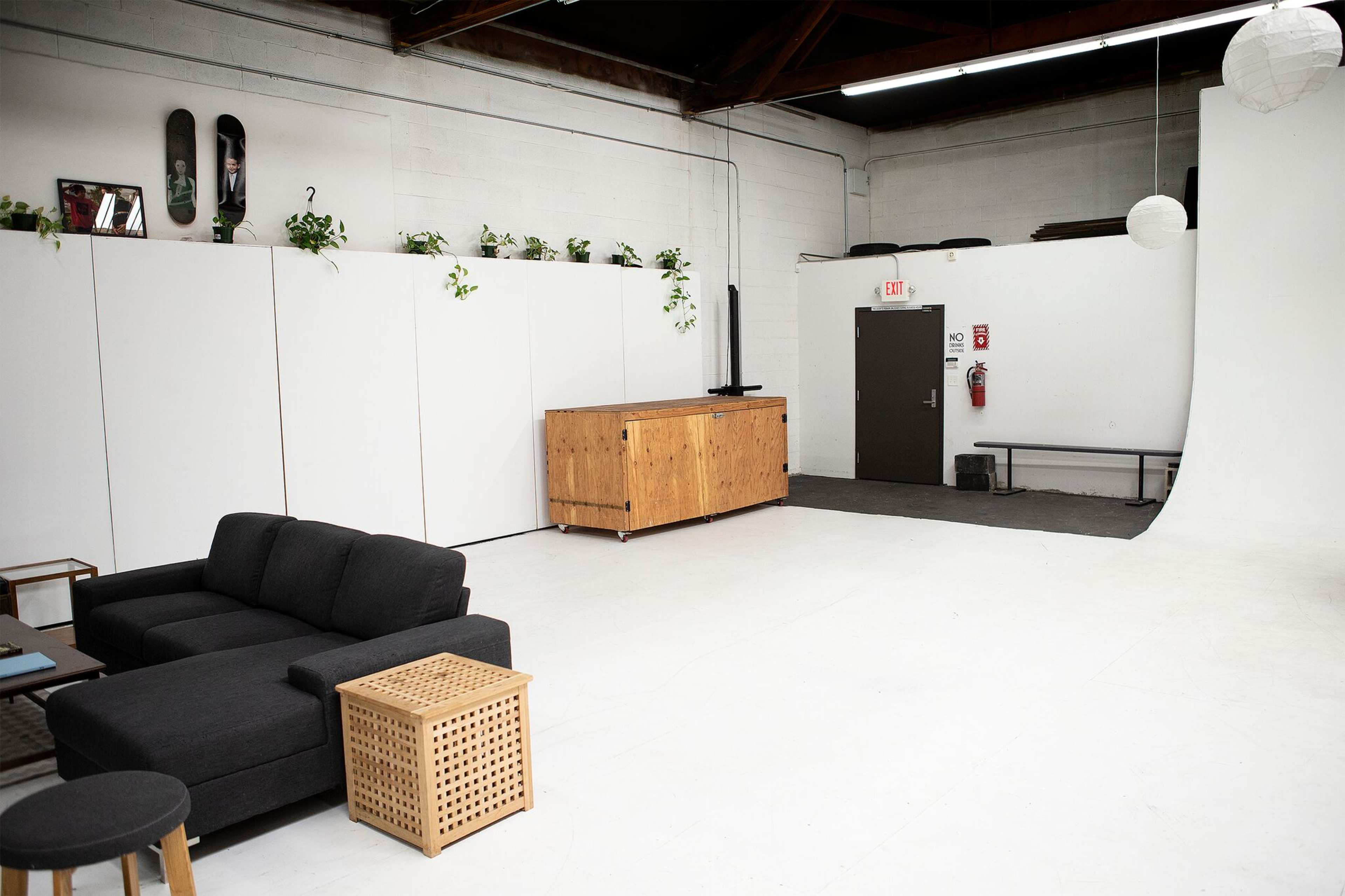 A spacious studio with a white backdrop, a wooden sideboard, a black sofa, and potted plants along the wall.