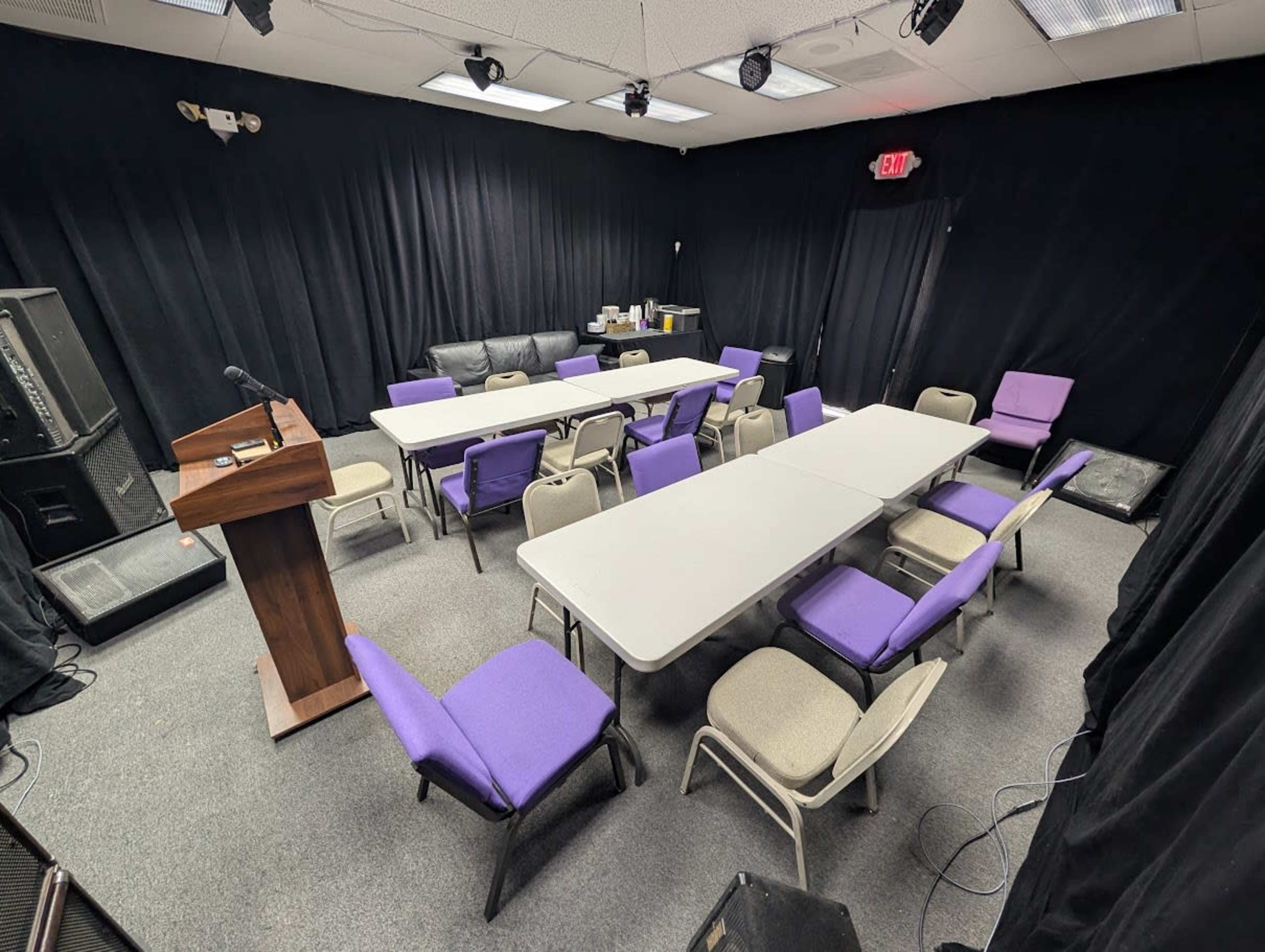 The image shows a room set up for a meeting, featuring several tables arranged in a U-shape, surrounded by chairs, with a podium in one corner and a dark backdrop.