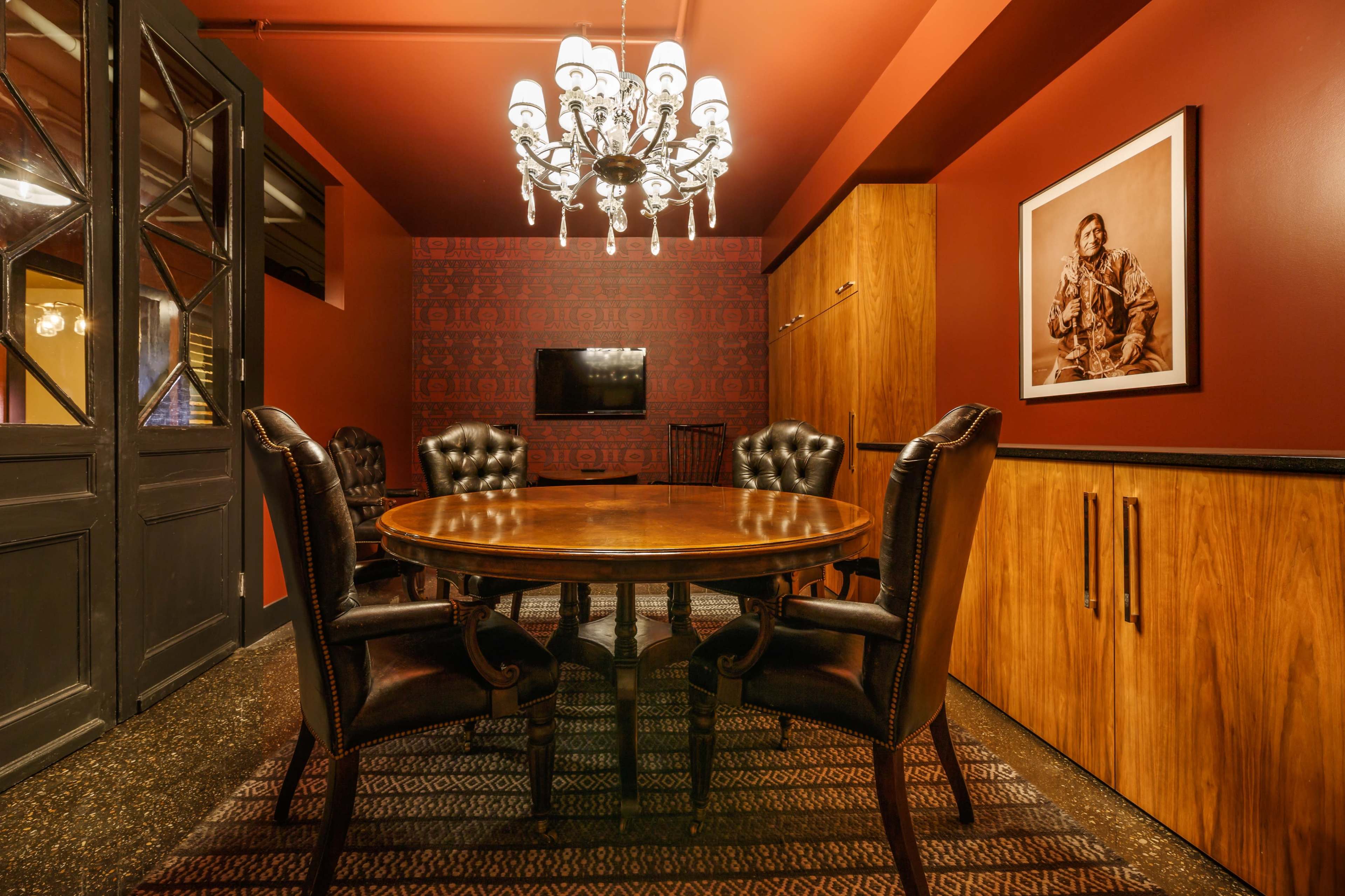 A dimly lit dining room with a circular wooden table surrounded by leather chairs, a chandelier overhead, and a television mounted on the wall.