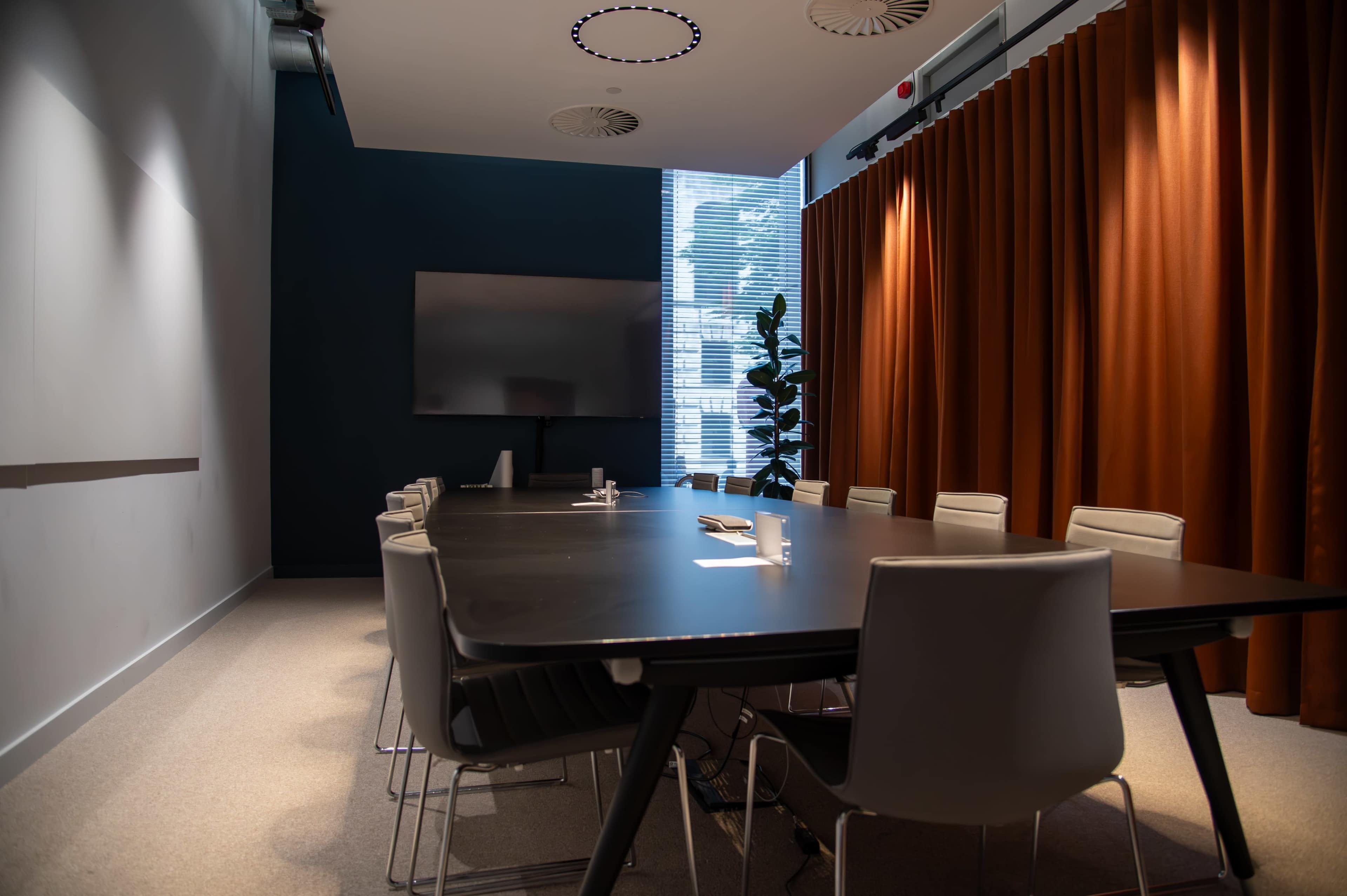 A modern conference room features a long table surrounded by chairs, with a large screen on one wall and curtains on the opposite side.