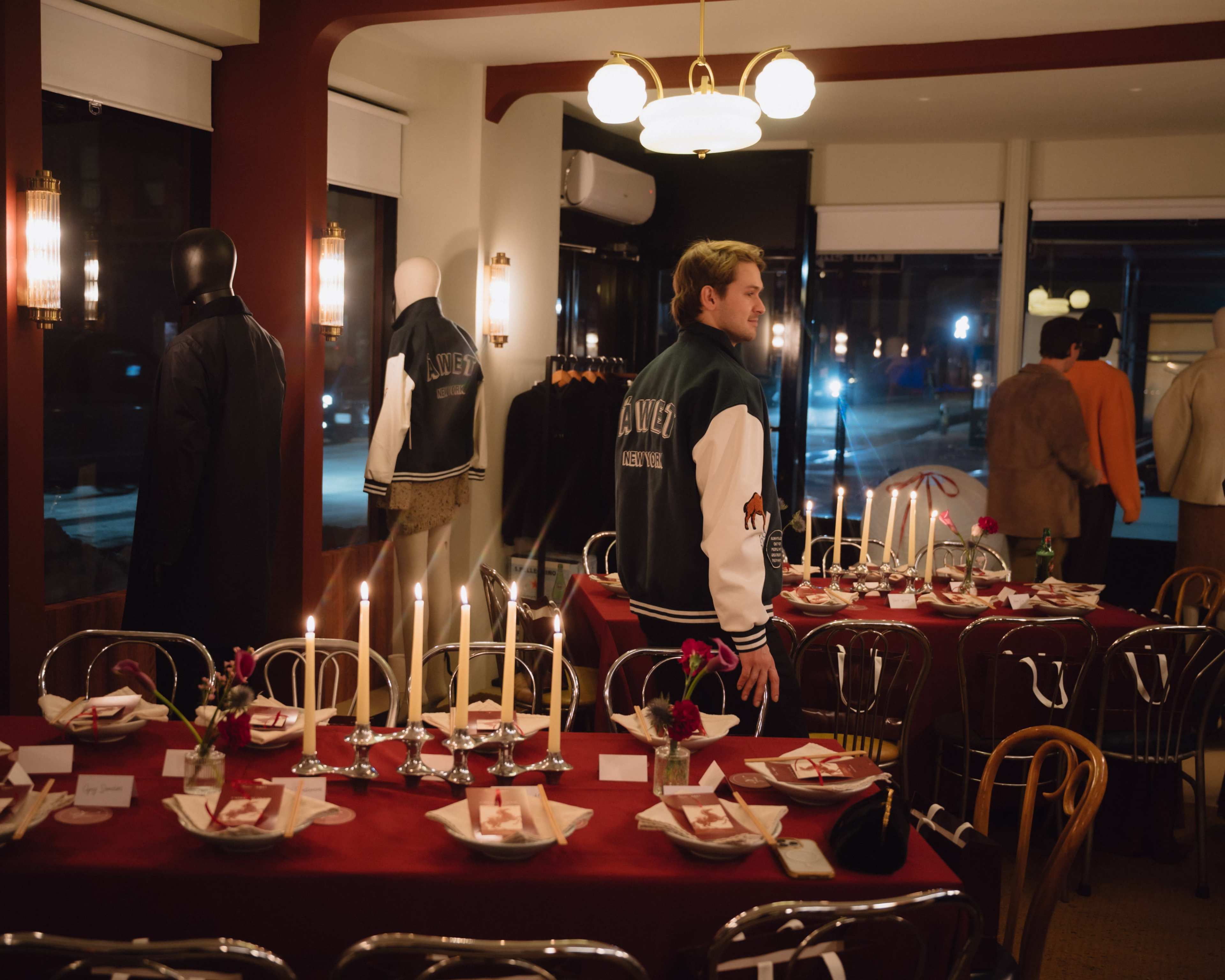 The scene shows a stylish dining setup with neatly arranged tables featuring candles and flowers, while mannequins dressed in jackets are positioned around the room.