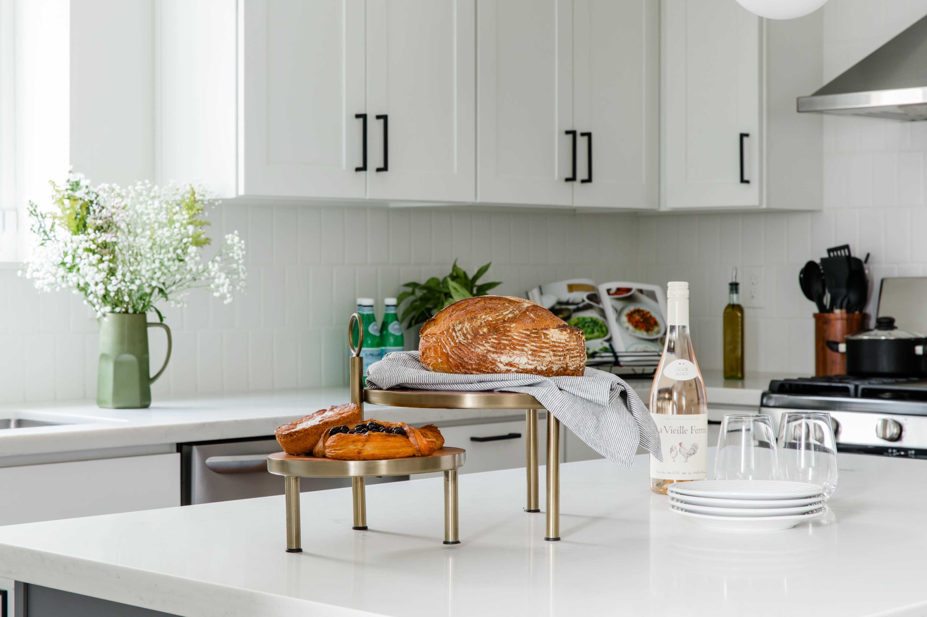 A wooden table displays a loaf of bread and pastries on a tiered stand, alongside a bottle of wine and plates in a modern kitchen.