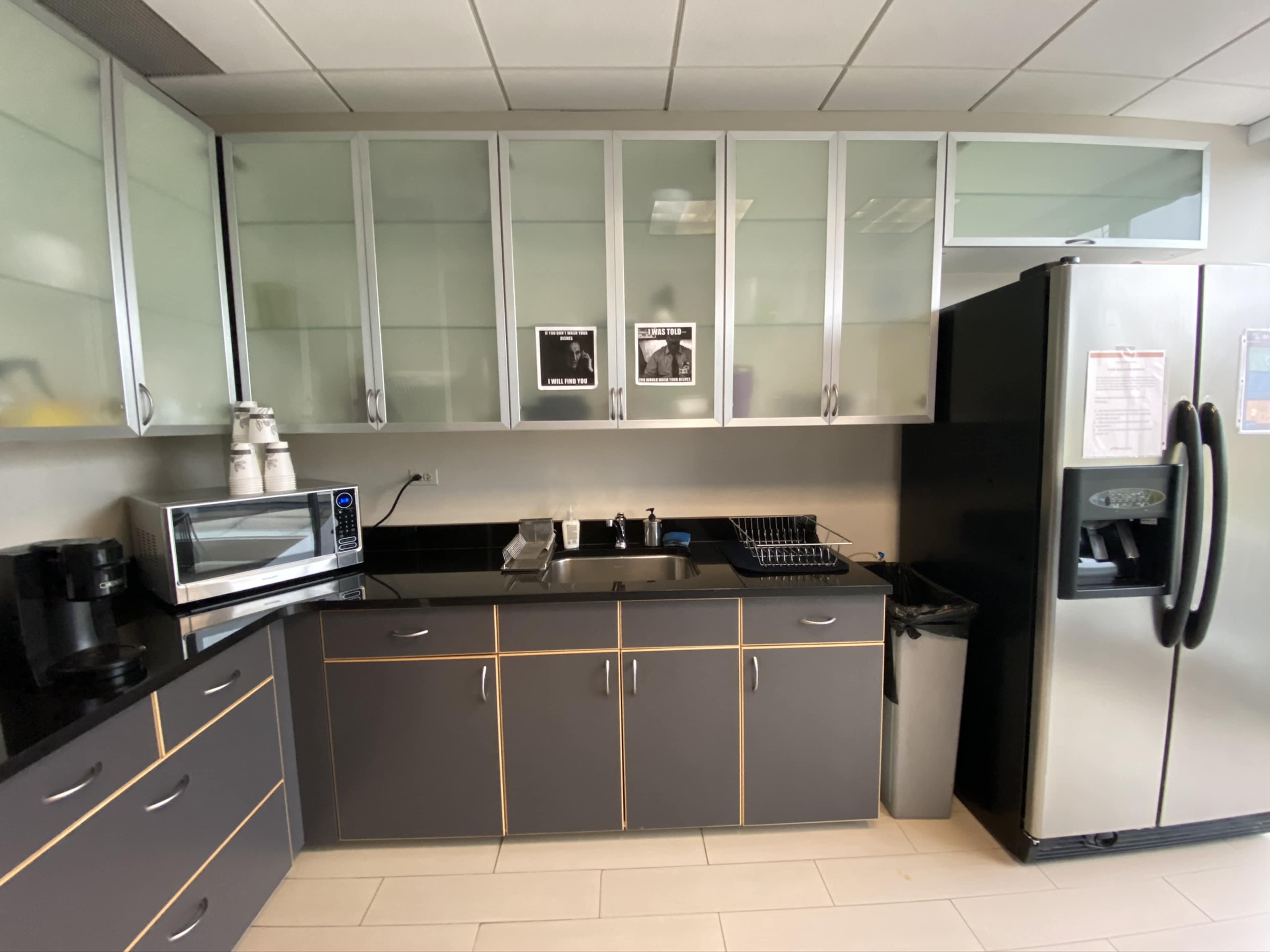 The image shows a modern kitchen with gray cabinets, a stainless steel refrigerator, a microwave, and a coffee maker, along with a dish rack and a trash bin on a black countertop.