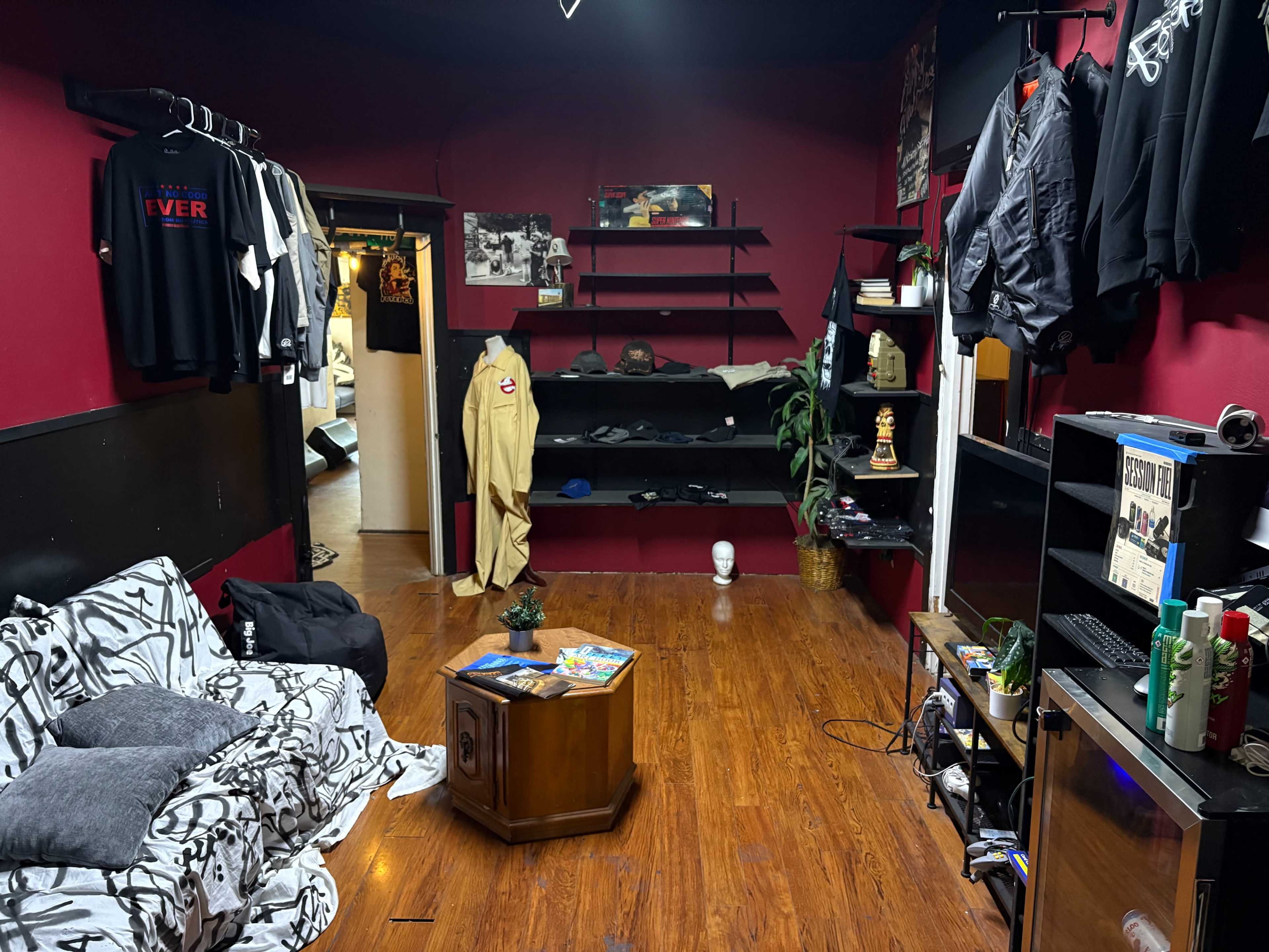 The image shows a room with red walls, wooden flooring, a couch covered in black and white fabric, and various clothing items hanging on shelves.