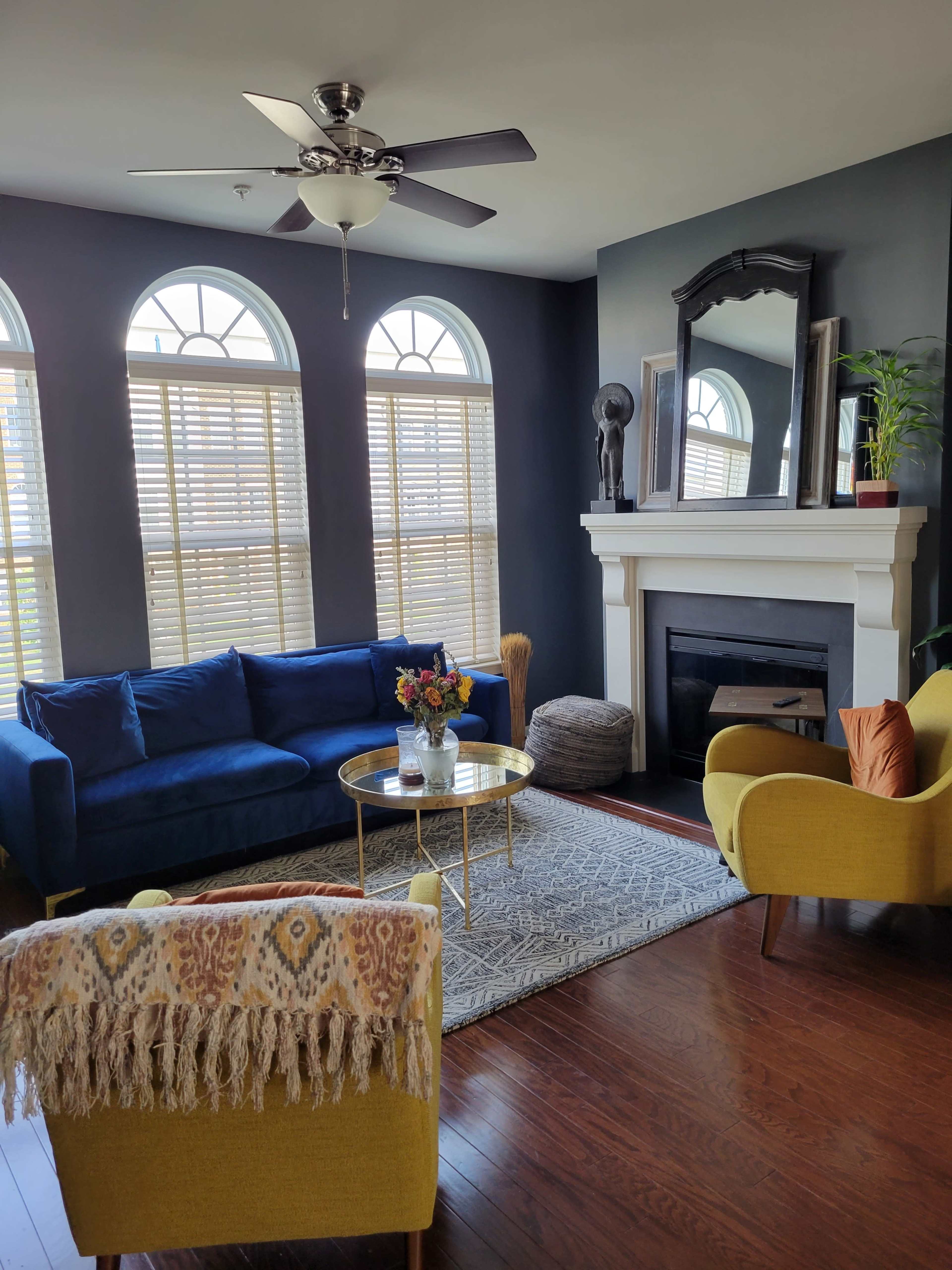 A living room features a blue sofa, yellow chairs, a round coffee table, and a fireplace beneath a large mirror, with natural light streaming through the arched windows.