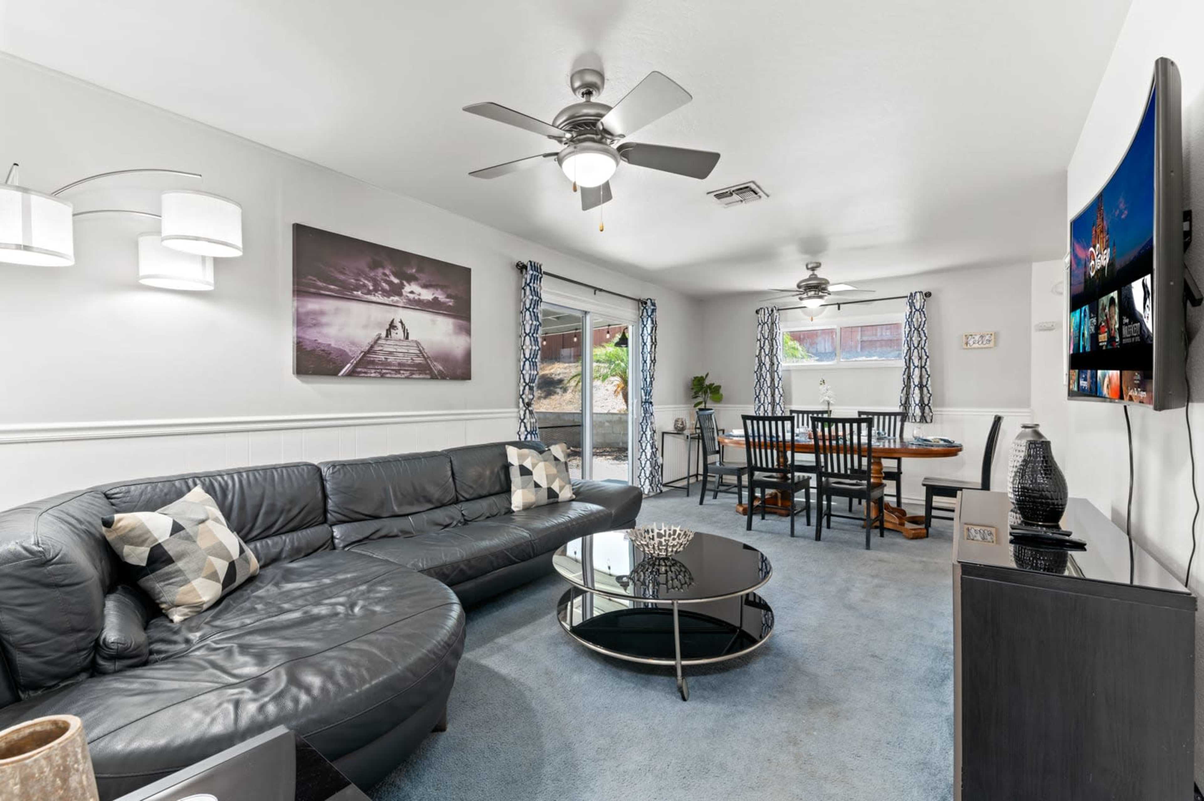 The image shows a living room featuring a black sectional sofa, a glass coffee table, a dining area with a wooden table and chairs, and large windows with blue curtains.