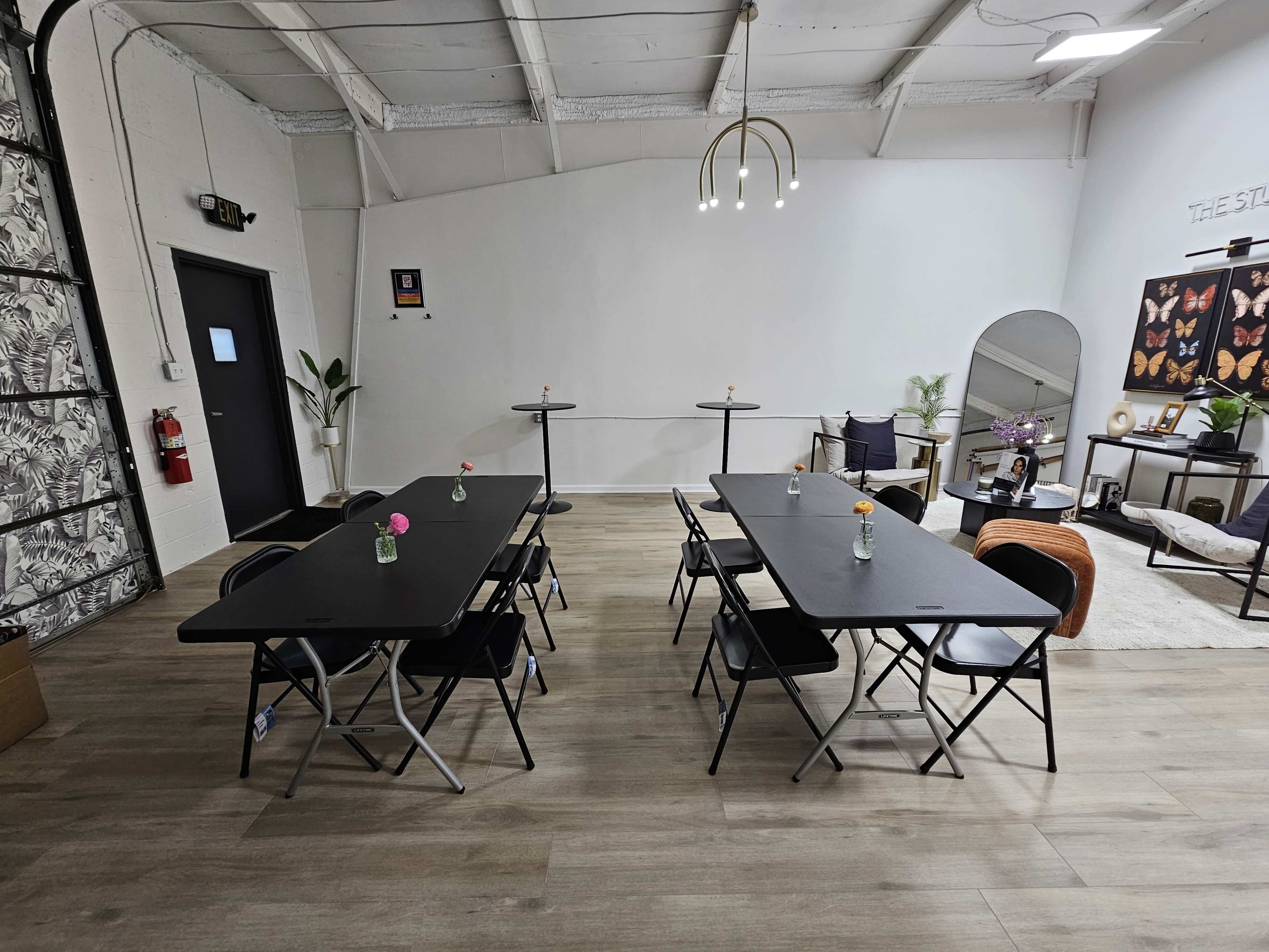 The image shows a spacious room with black folding tables and chairs arranged neatly, a small lounge area with a couch, and a decorative wall featuring butterfly art.