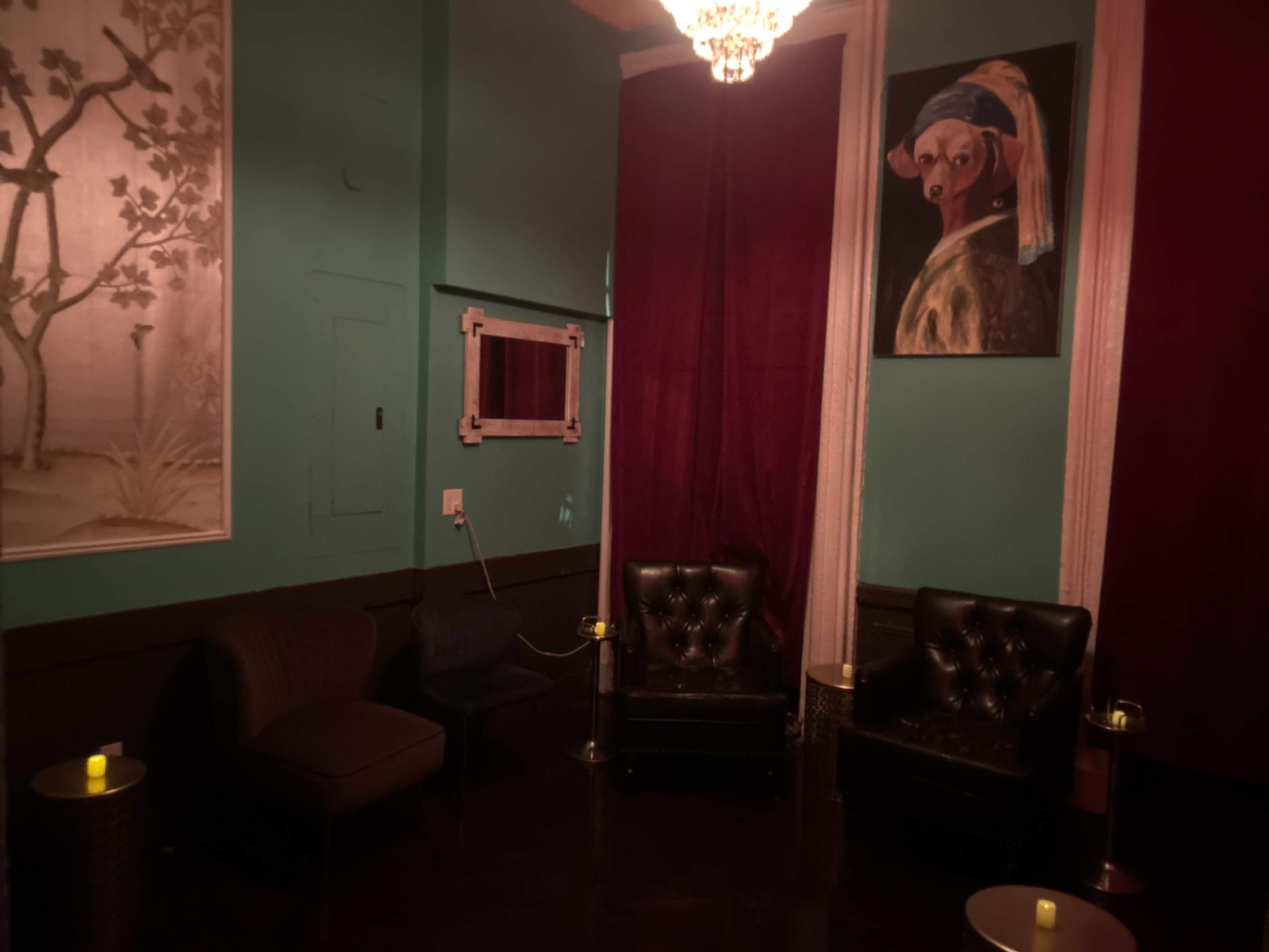 The image shows a dimly lit lounge area featuring two black leather chairs, a small side table with candles, and a wall adorned with a large painting and red curtains.