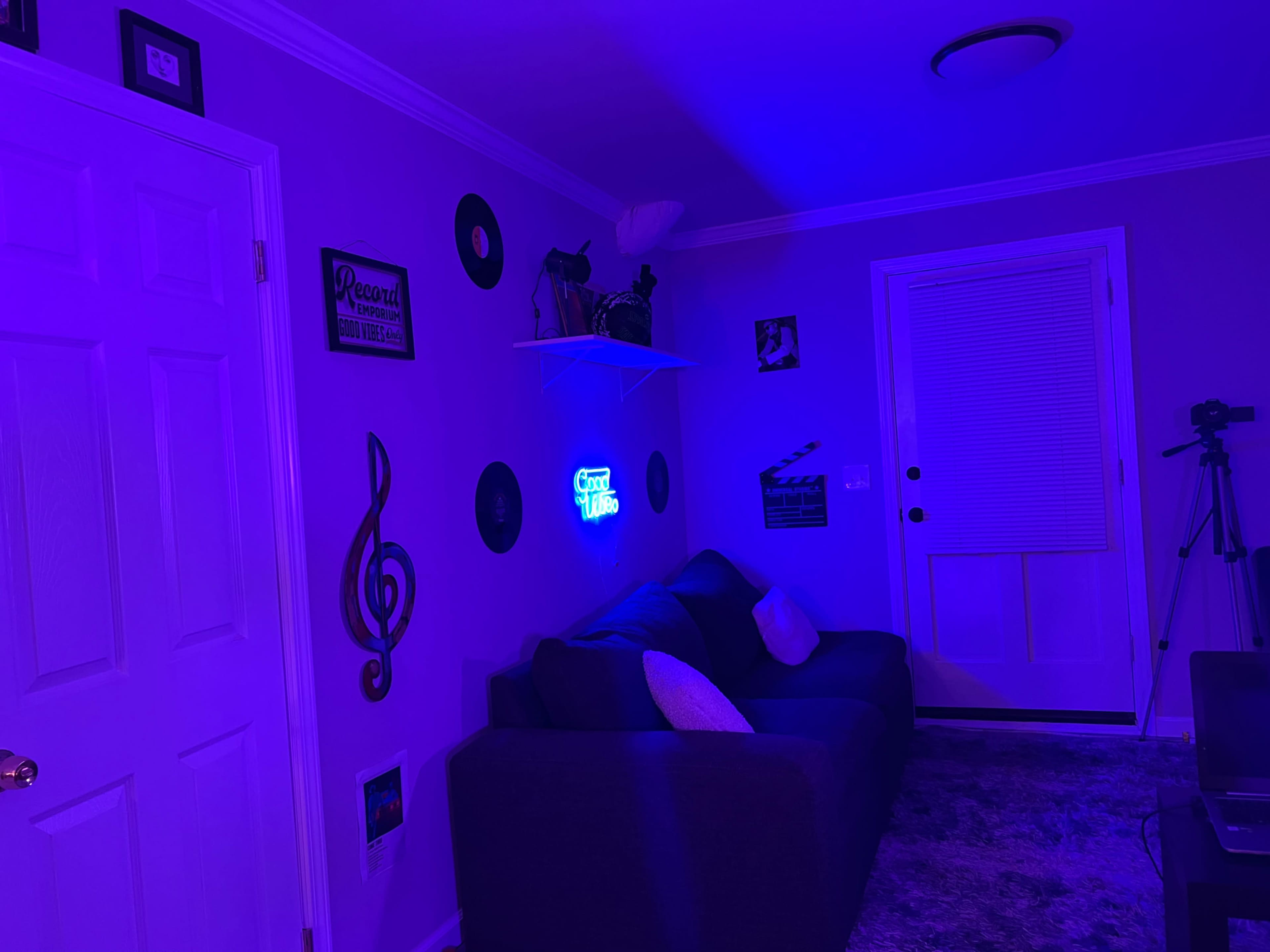 A dark room features a sofa with white pillows, purple walls, and decorations including records, musical notes, and a glowing neon sign.