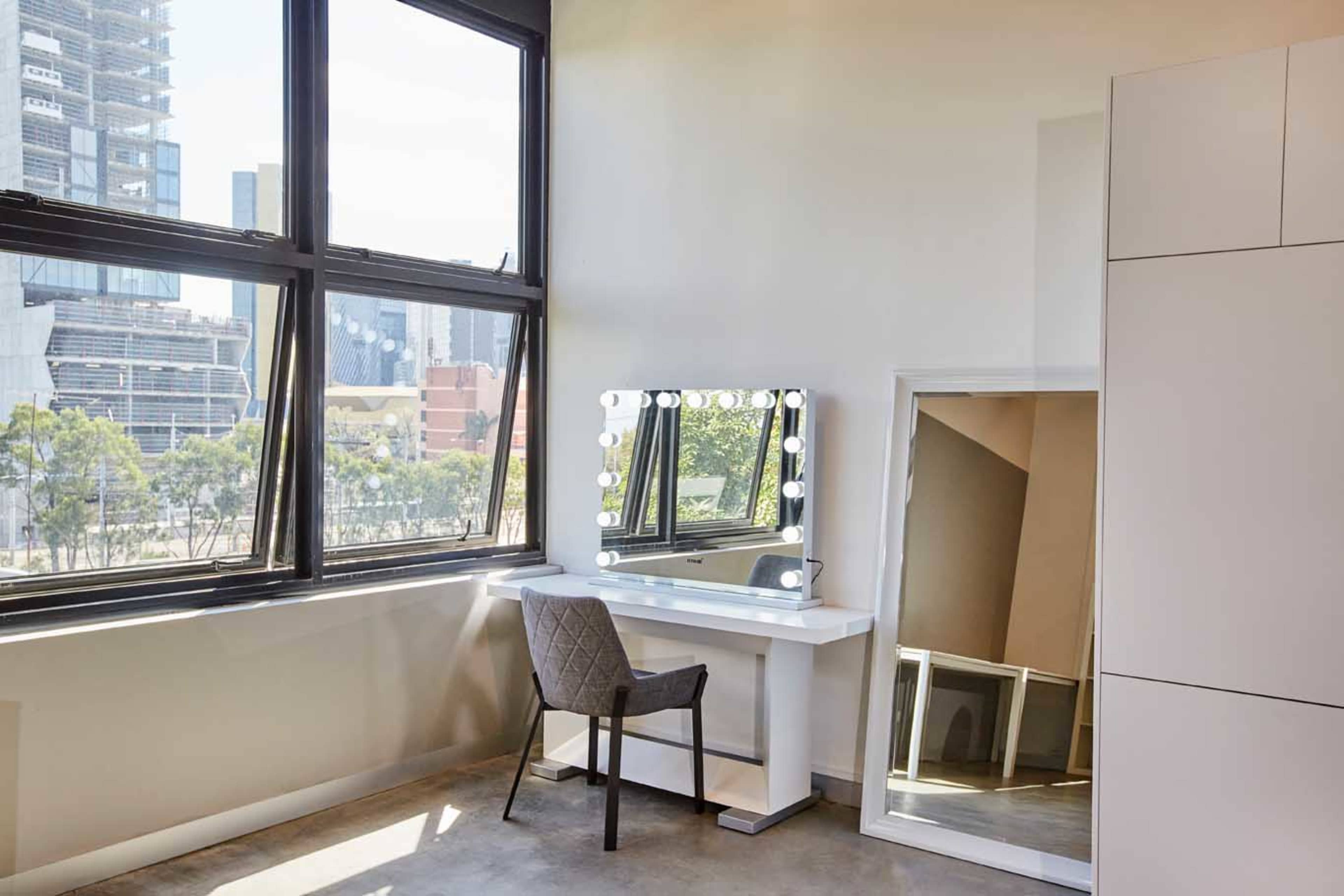 A modern vanity area with a lighted mirror and a chair is located beside large windows in a room with a view of a city skyline.