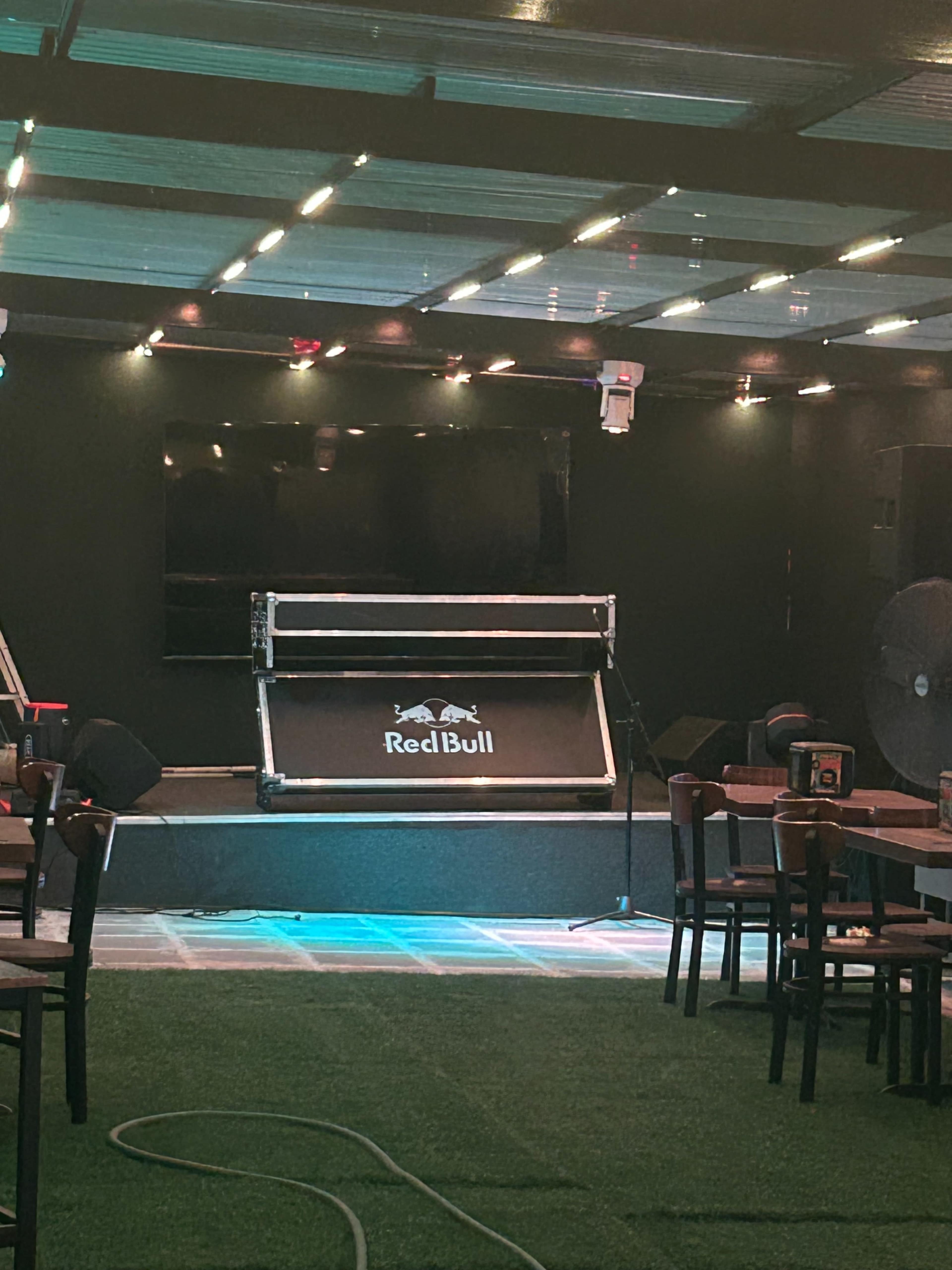 The image shows a dimly lit stage area in a venue featuring a microphone stand, a backdrop with the Red Bull logo, and rows of wooden chairs set up for an audience.