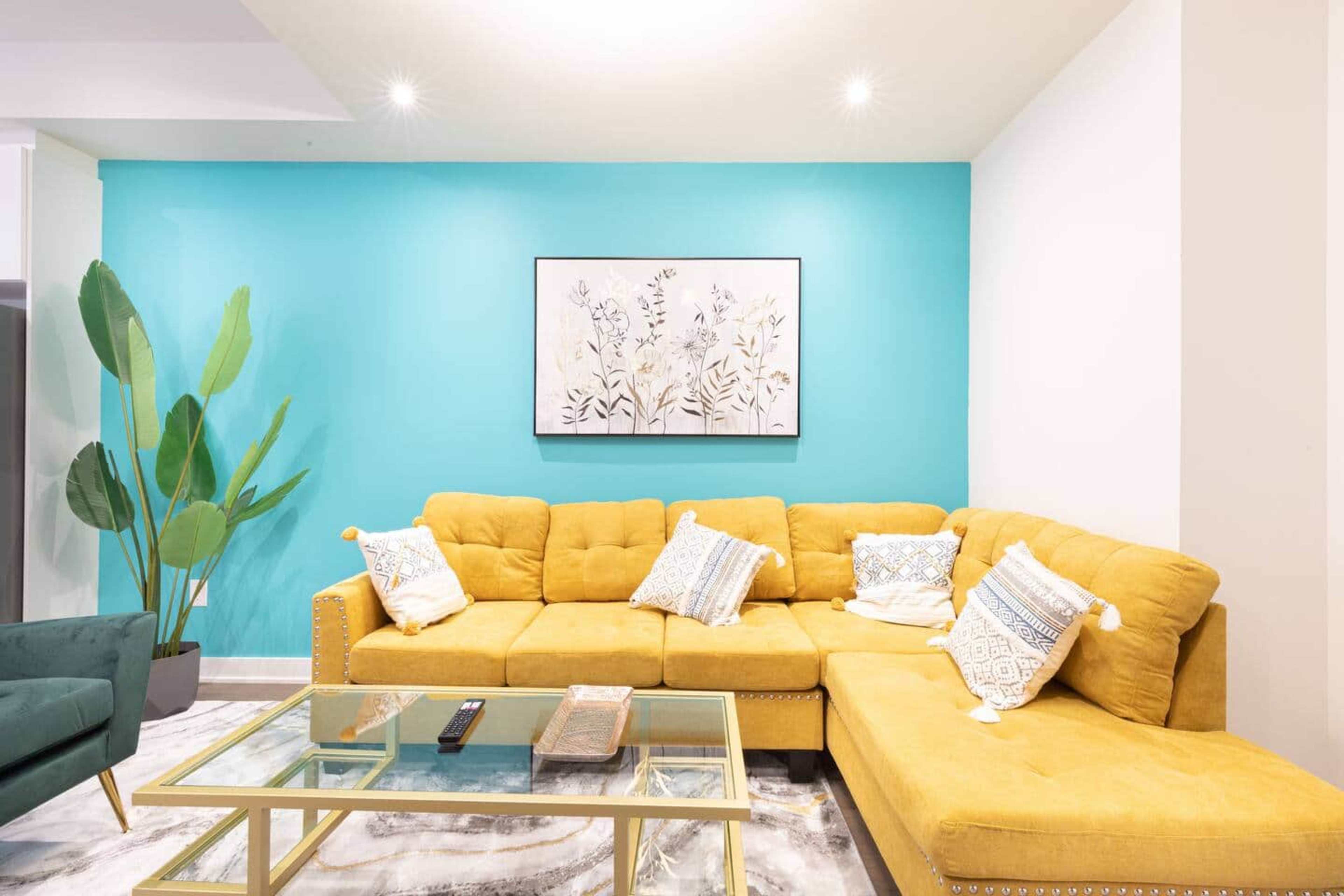 The image features a modern living room with a yellow sectional sofa, a glass coffee table, and a vibrant teal accent wall adorned with a framed floral artwork.