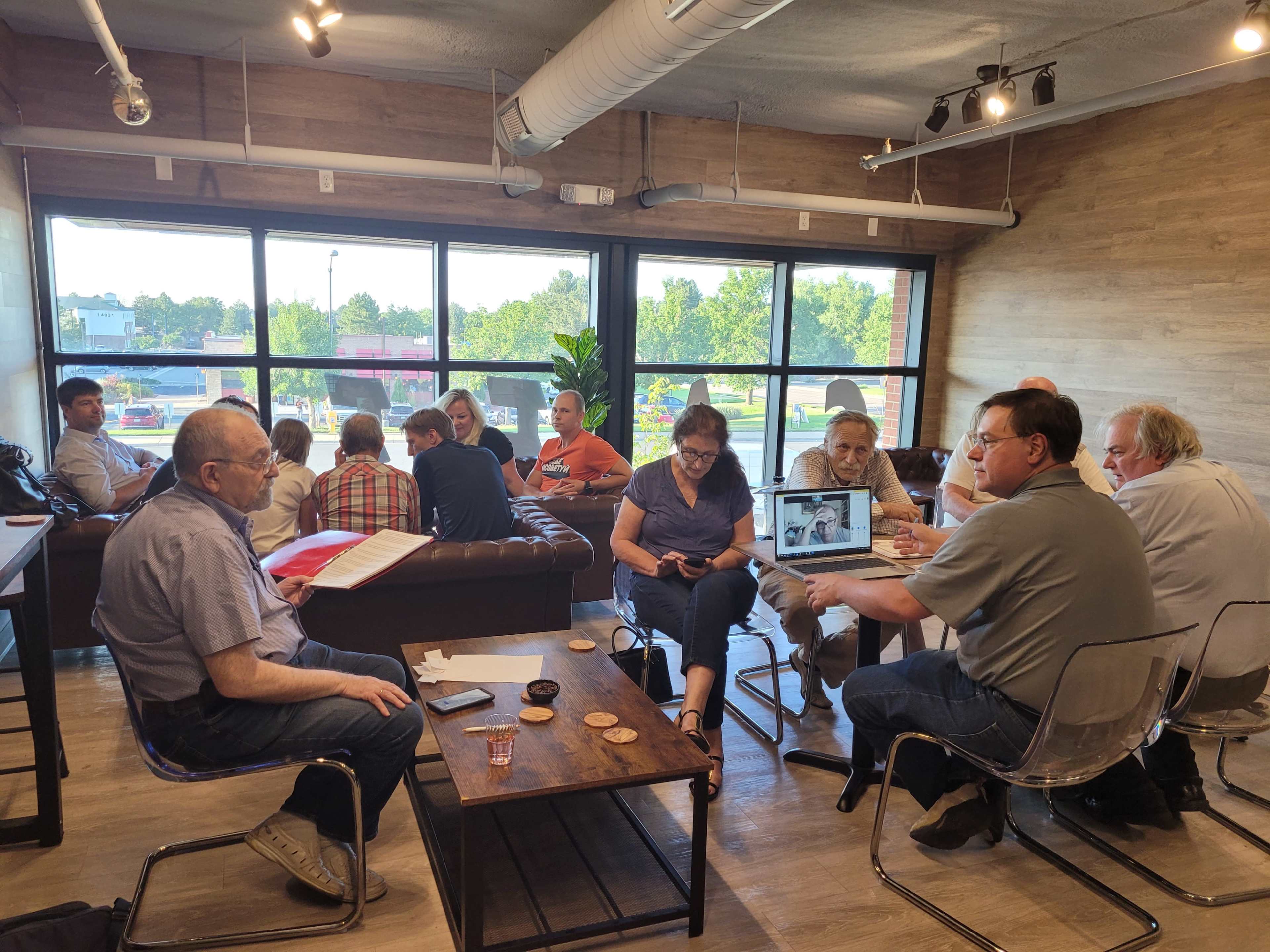 A group of people is gathered in a well-lit café, engaged in conversation and using laptops around wooden tables.