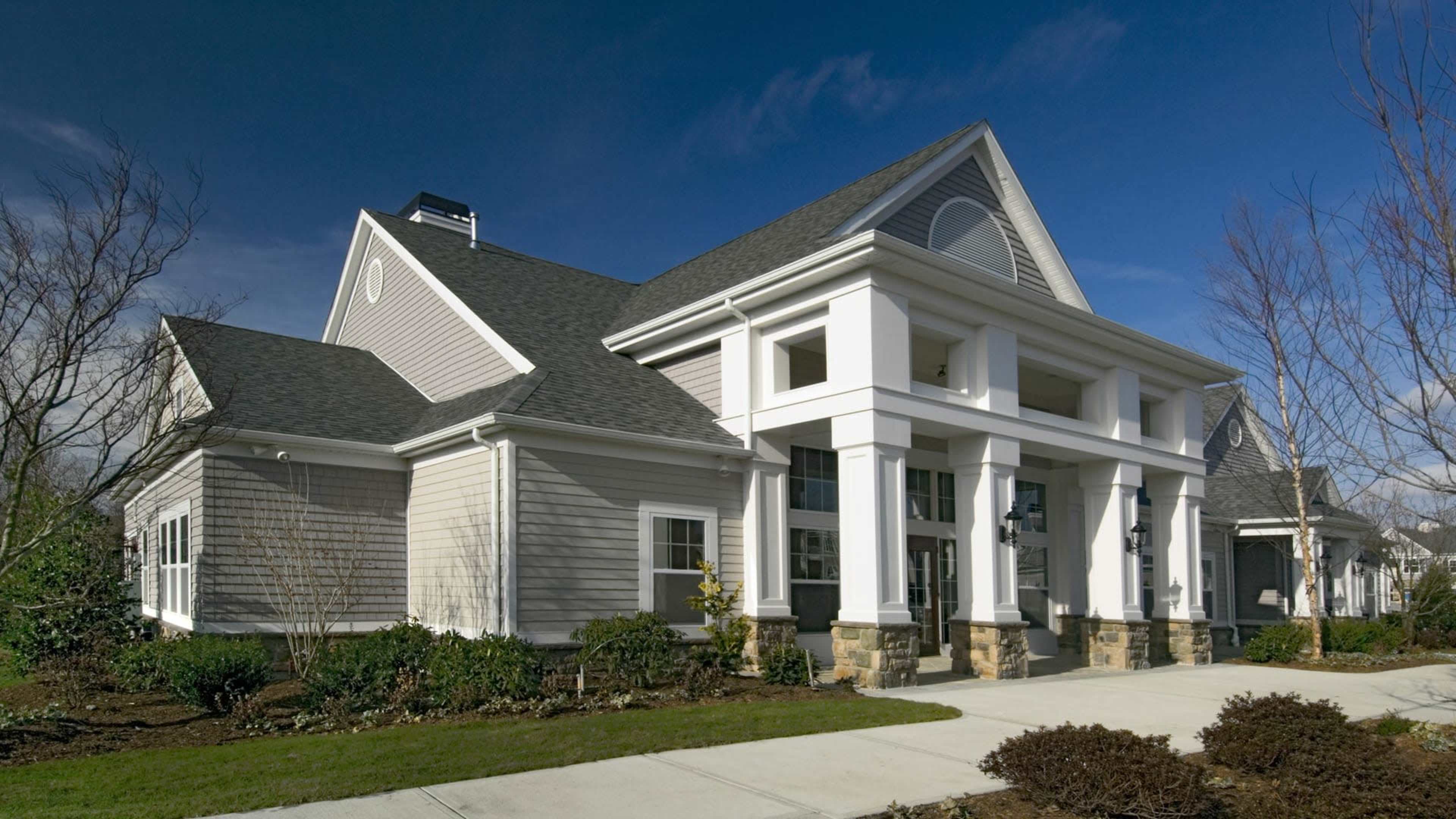 The image shows a modern, single-story building with a gabled roof, large front columns, and a landscaped entrance path.