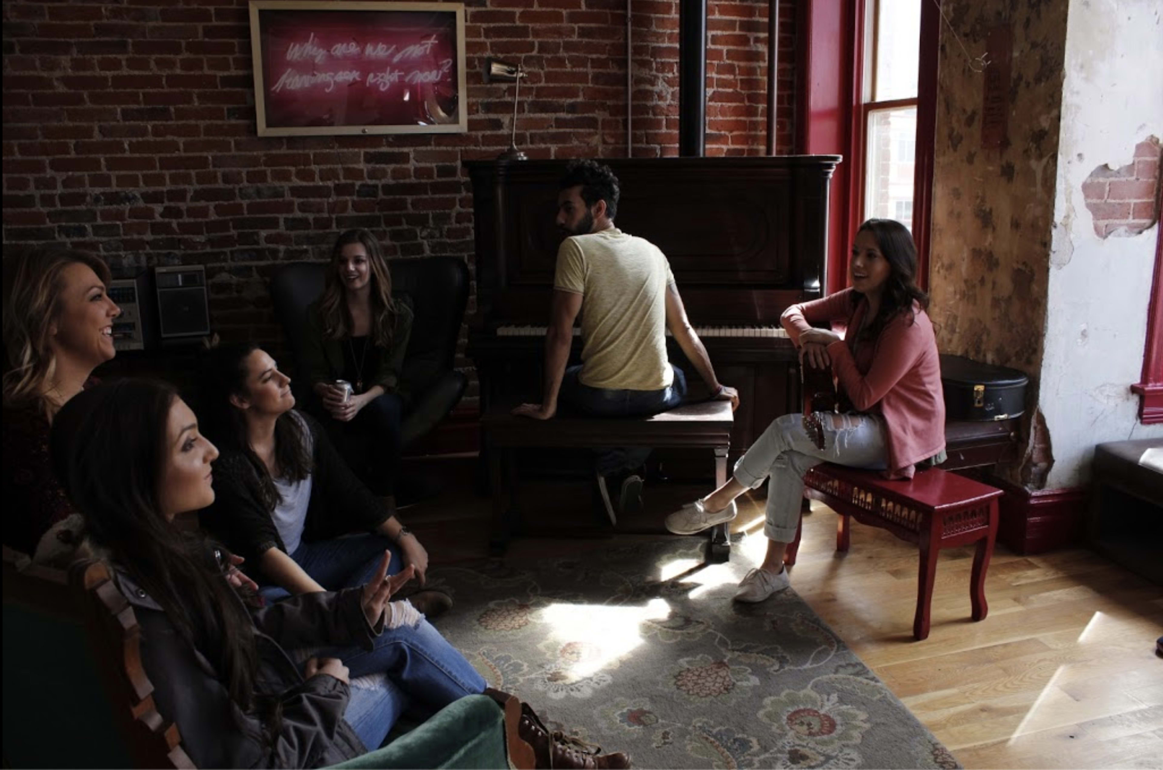 A group of six people sits in a cozy room with exposed brick walls, while one person plays the piano.
