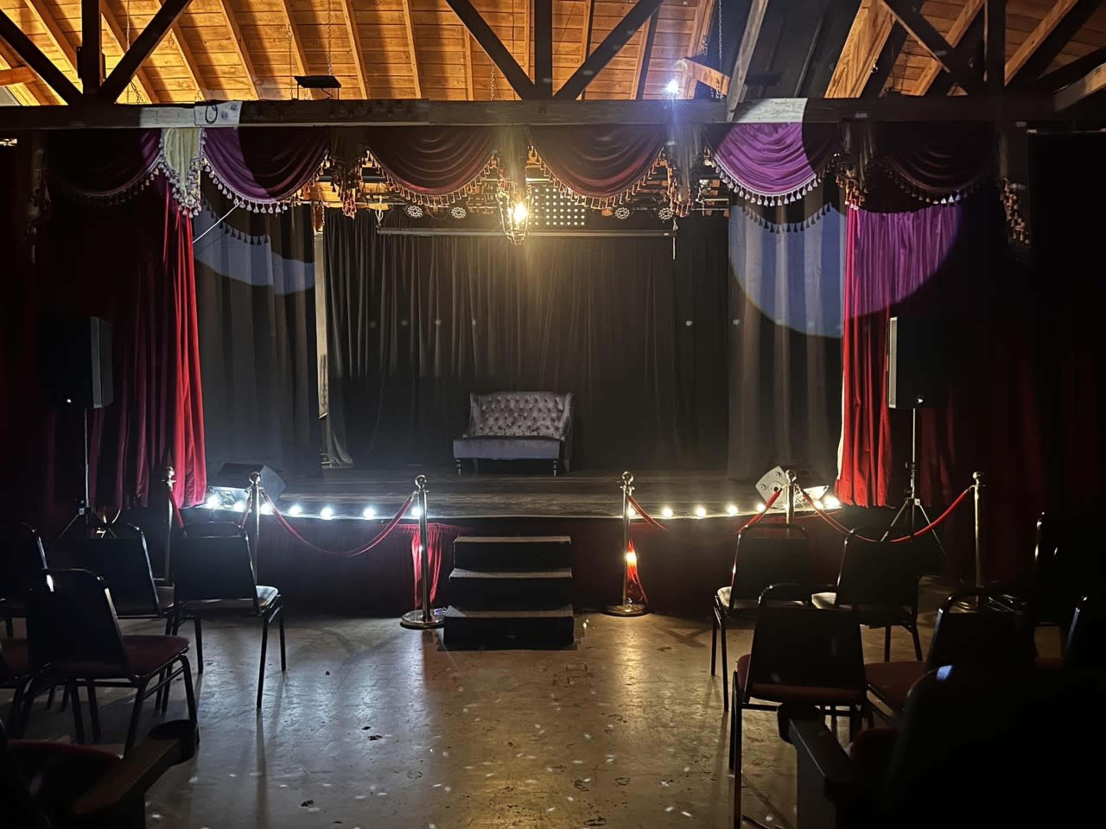 A small stage is set with a lone couch and illuminated by spotlights, surrounded by empty chairs and red velvet curtains.