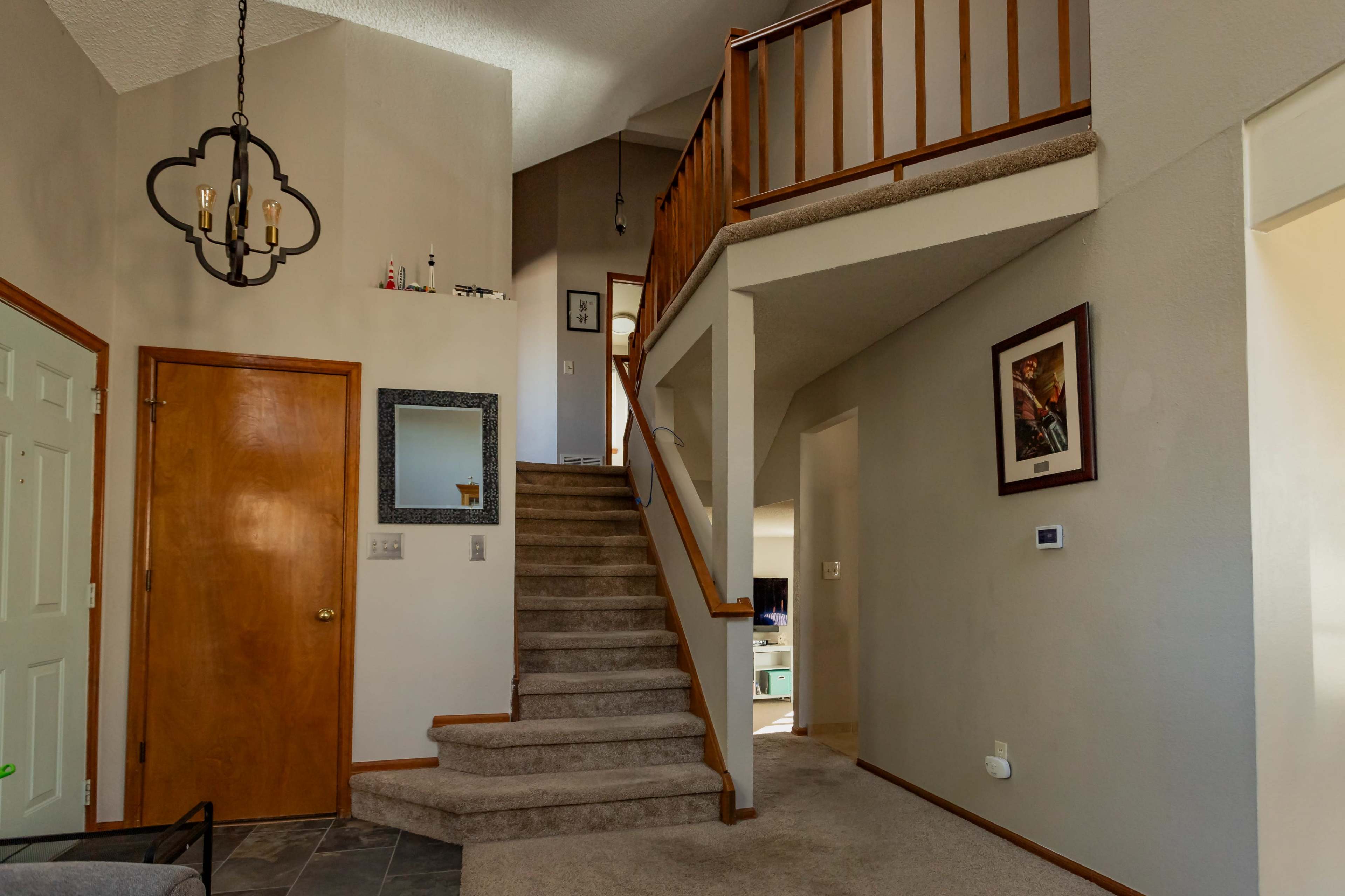 The image shows a well-lit entryway with a staircase leading to a second floor, featuring a chandelier and framed artwork on the walls.