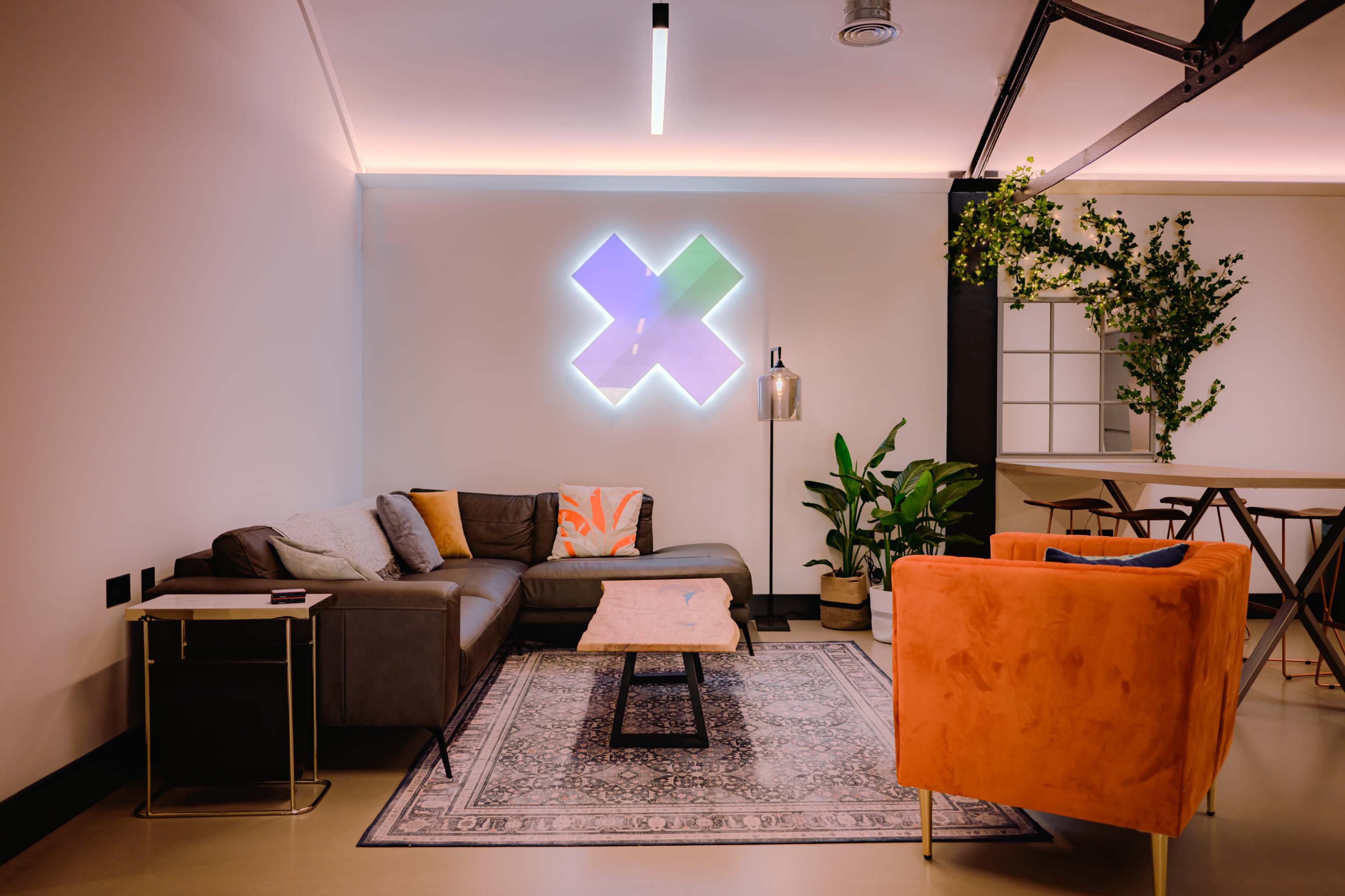 A modern lounge area features a sectional sofa, an orange armchair, a wooden coffee table, and decorative plants against a backdrop of geometric wall art.