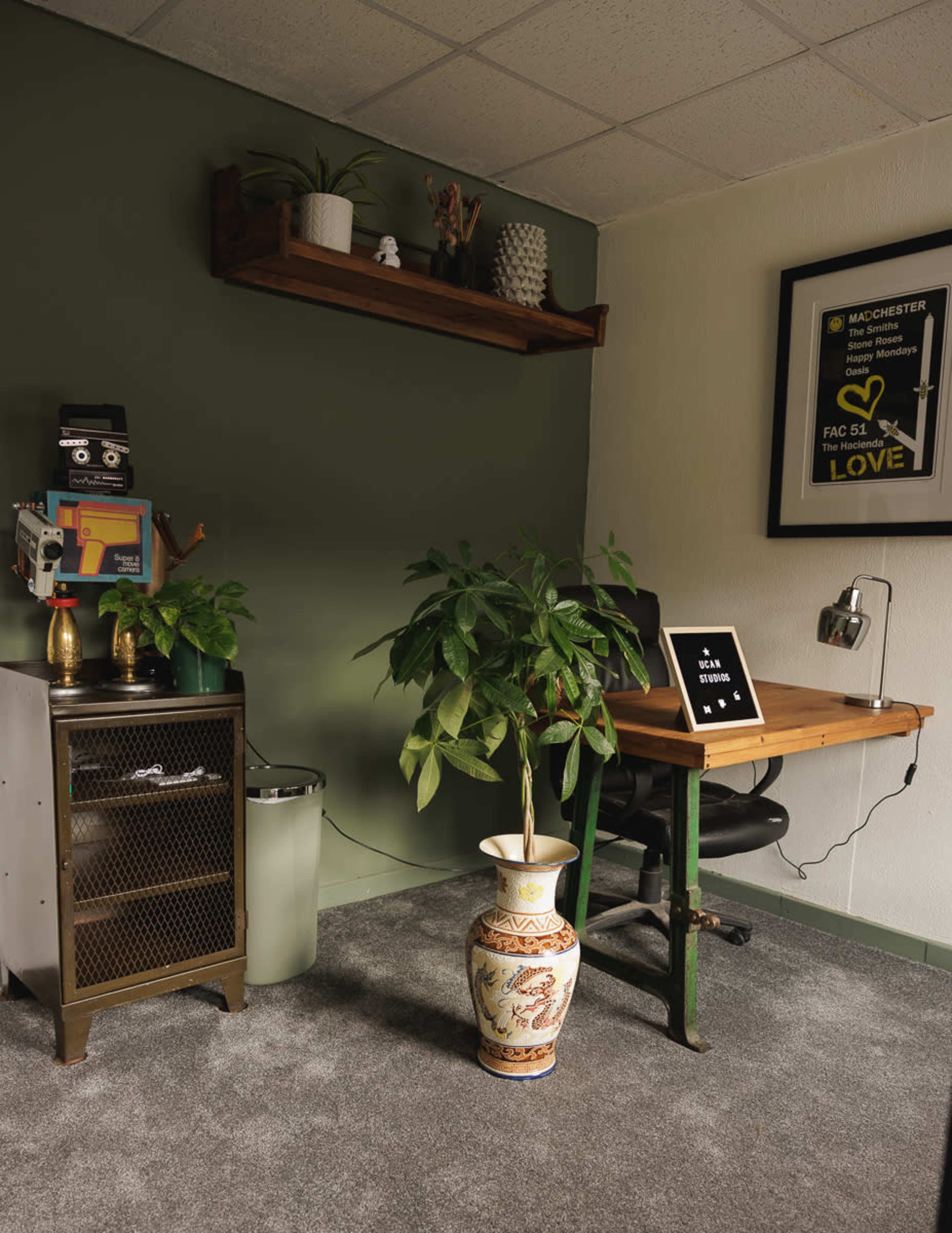 The image shows a small office space with a desk, a potted plant, a vintage filing cabinet, and shelves displaying decor items on a green wall.