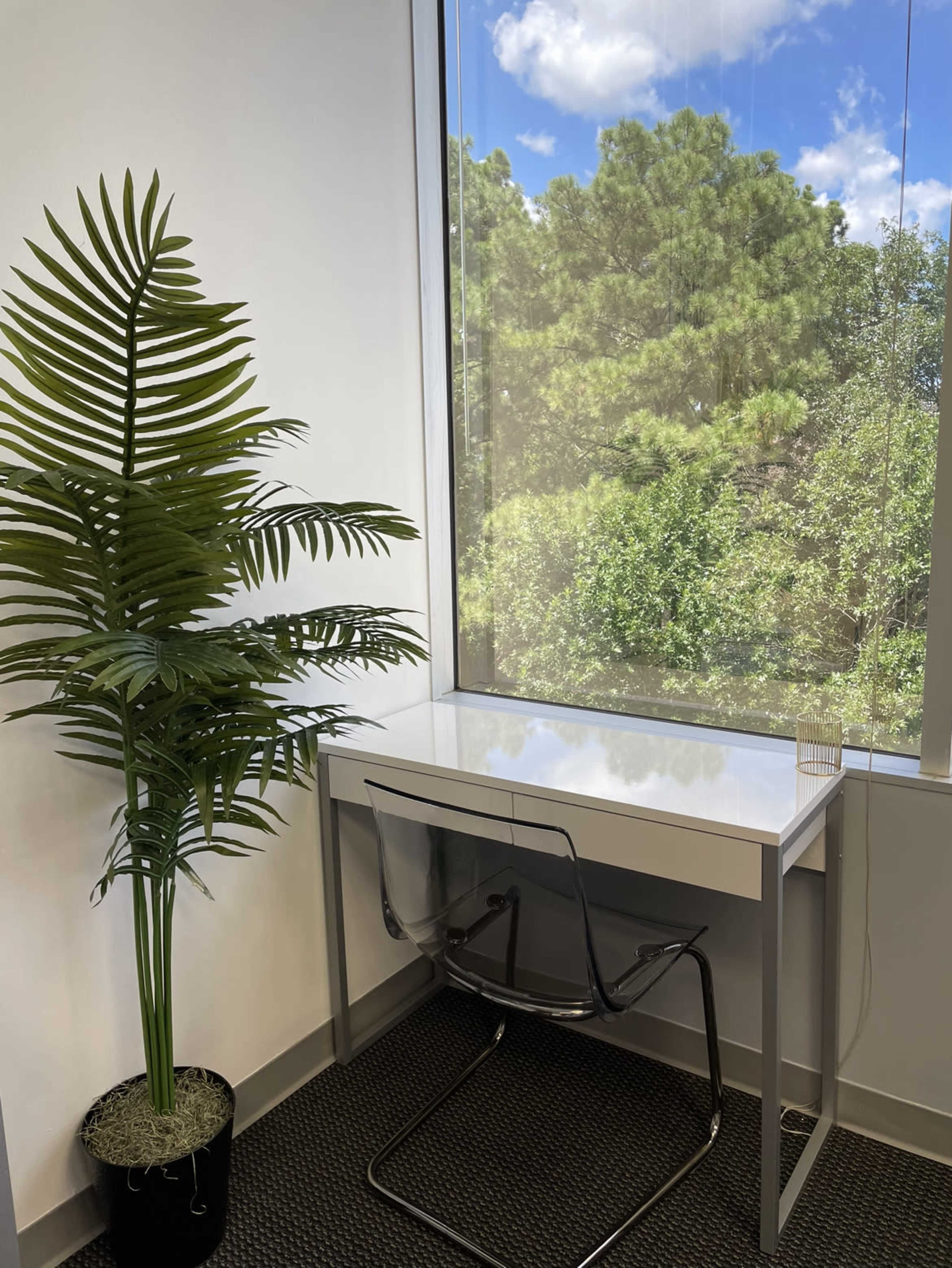 The scene features a modern desk with a clear chair beside a large window showcasing a green landscape and a potted palm plant.