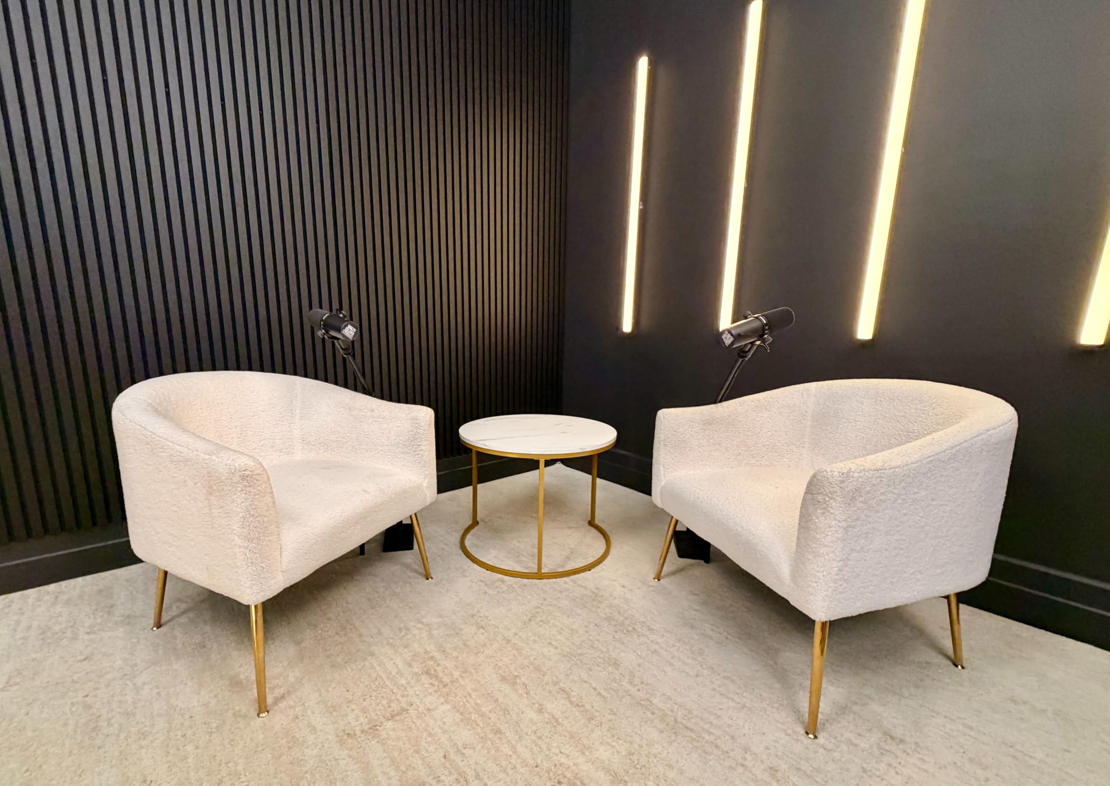 The image shows two modern light-colored armchairs positioned around a small round table with a gold base, set against a black paneled wall with vertical lighting.