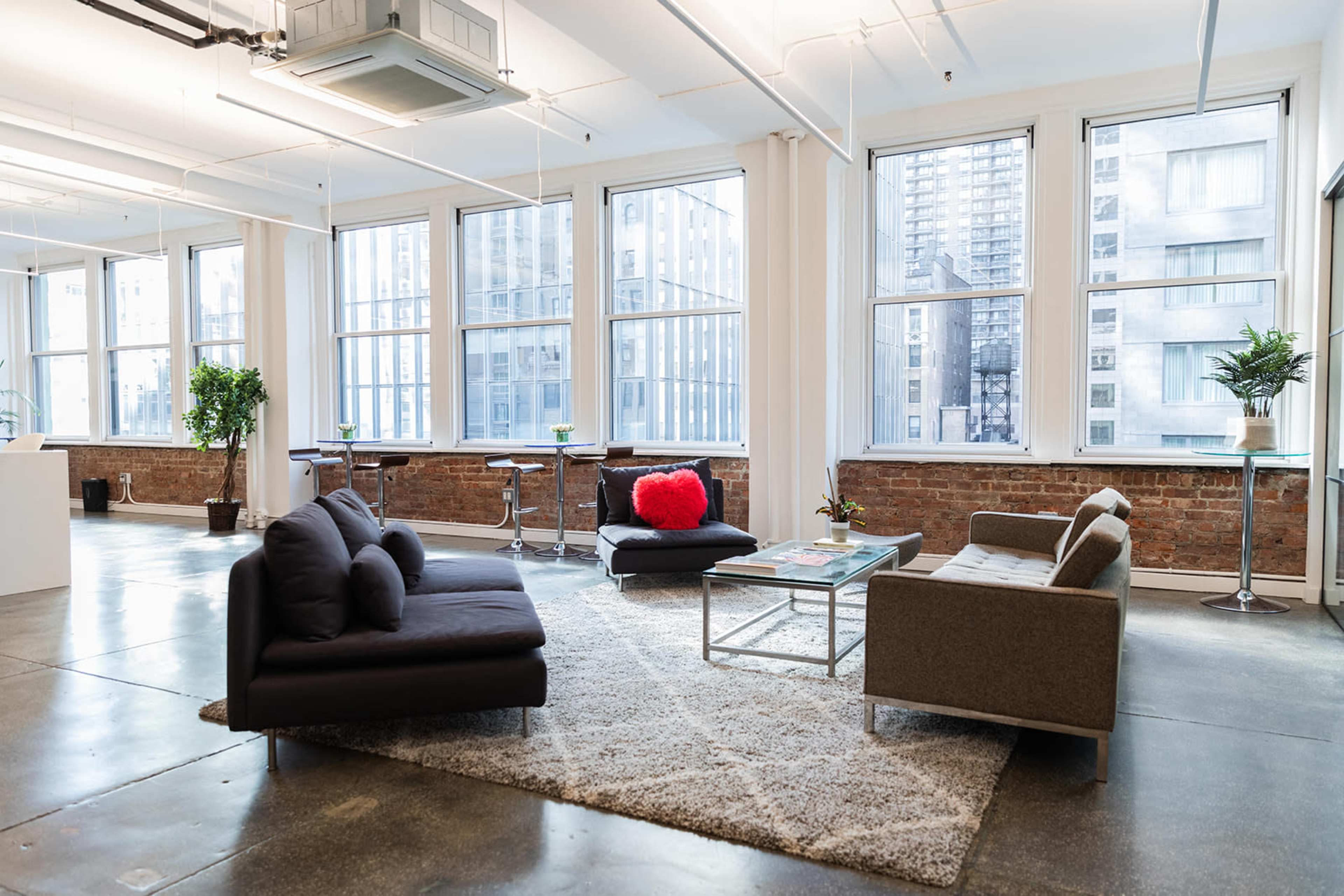 A modern office space featuring large windows, a gray sofa, a beige couch, and a coffee table on a carpet, with greenery in the corners.