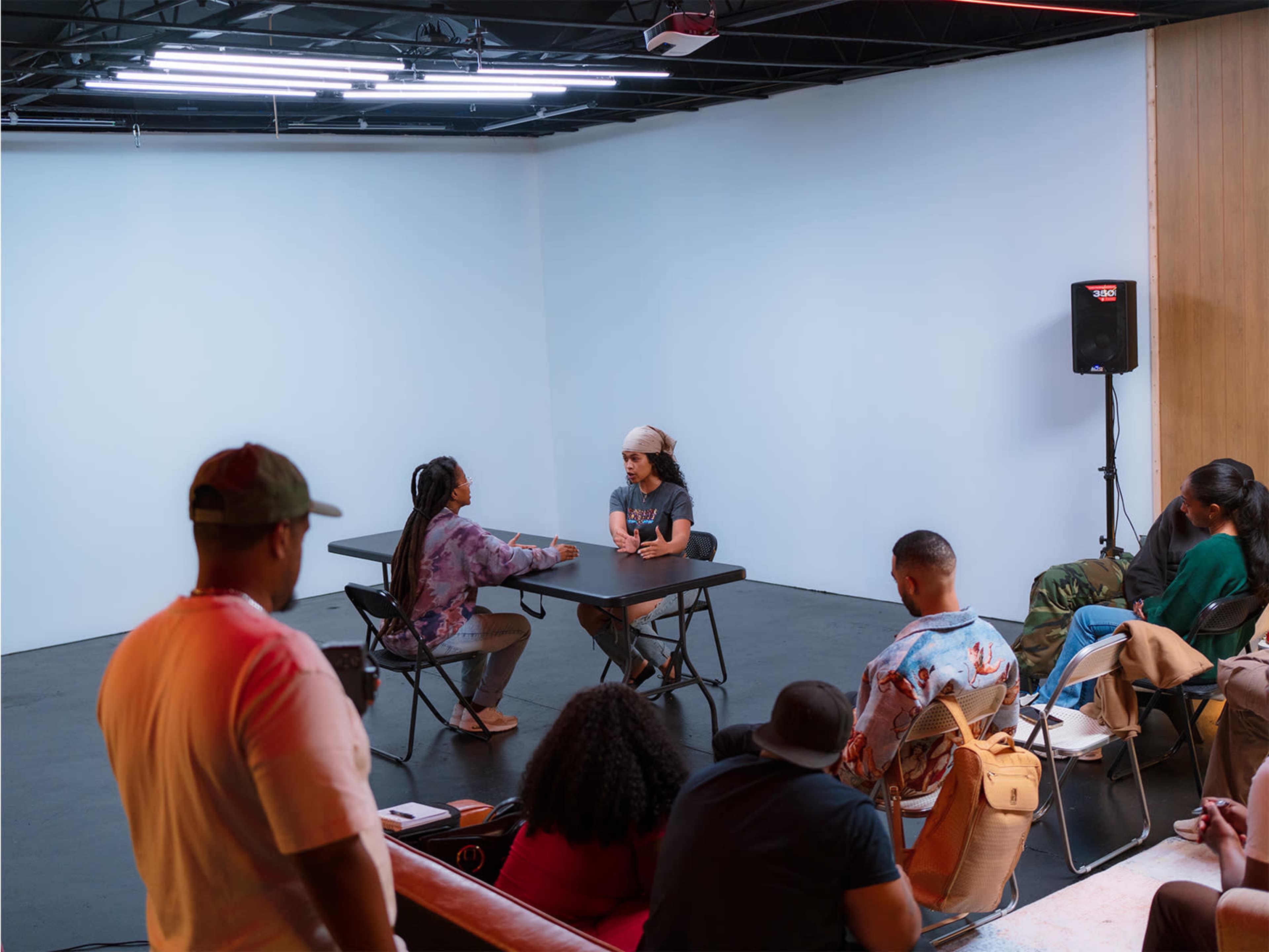 Two people are seated at a table engaged in conversation, while a small audience watches from a distance in a minimalist indoor space.