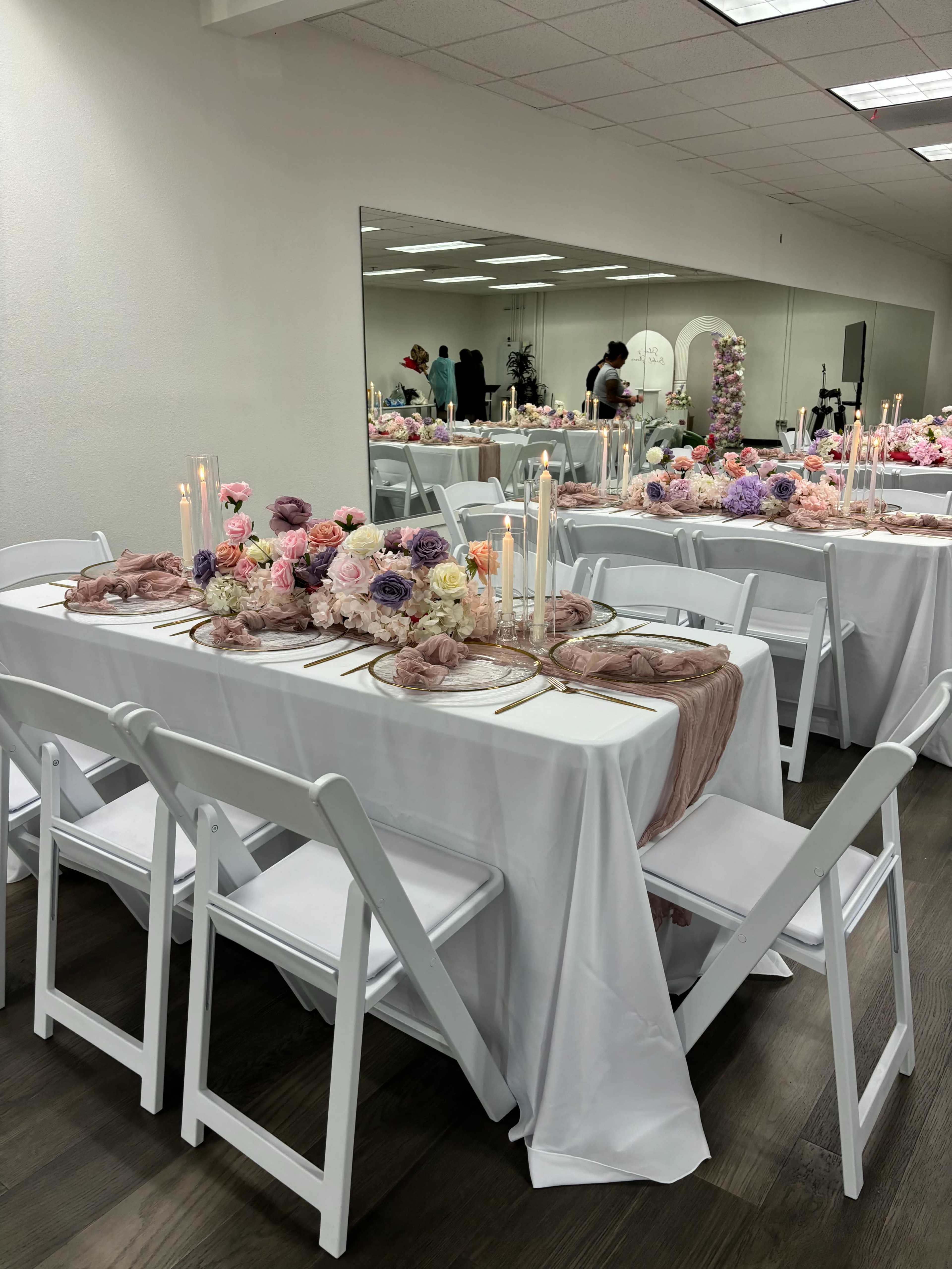 The image shows a banquet setup with long tables adorned with floral centerpieces and candles, surrounded by white chairs in a well-lit room.