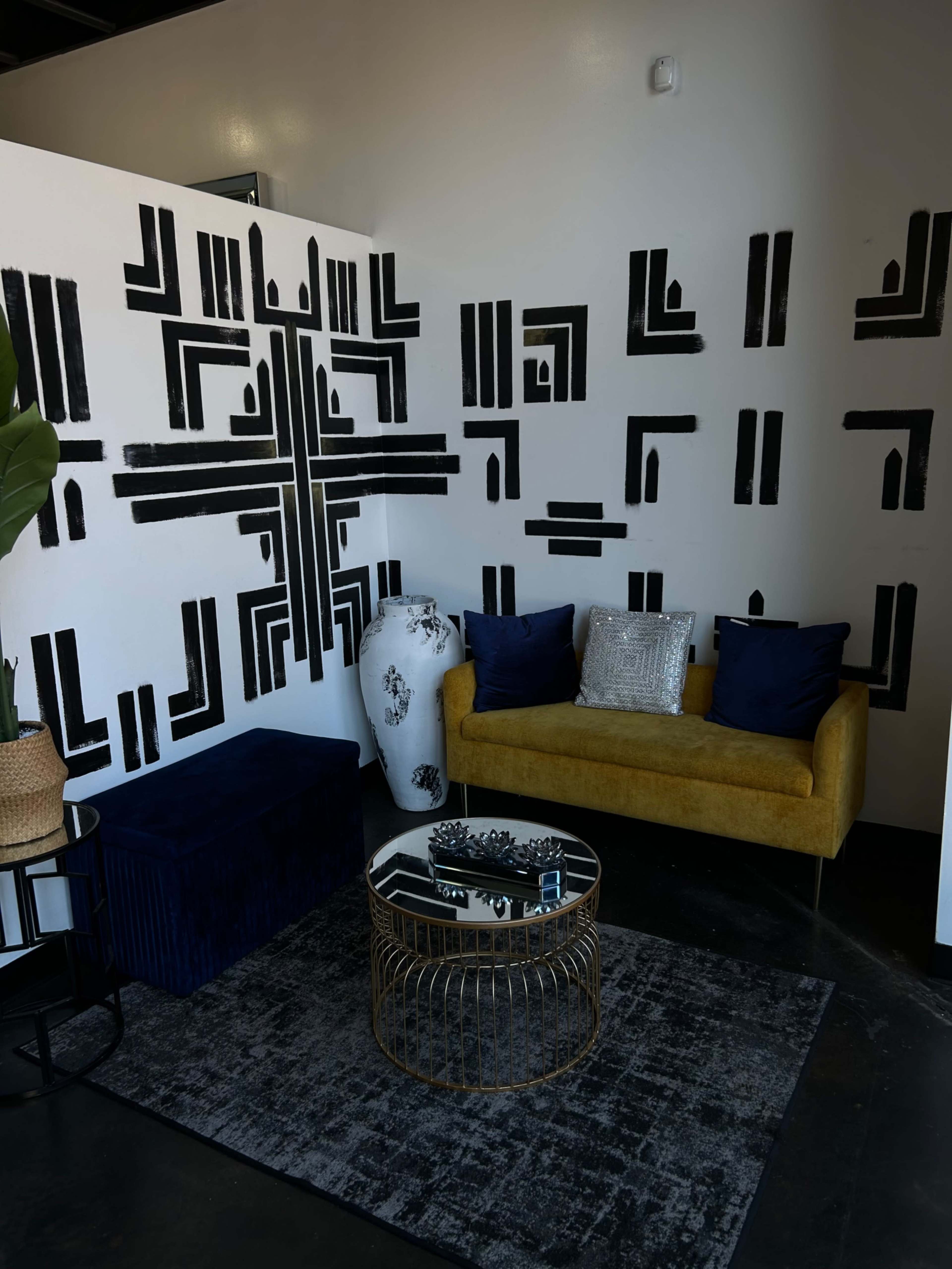 A modern seating area with a yellow sofa, a black patterned wall, and decorative elements including a glass coffee table and a large plant.
