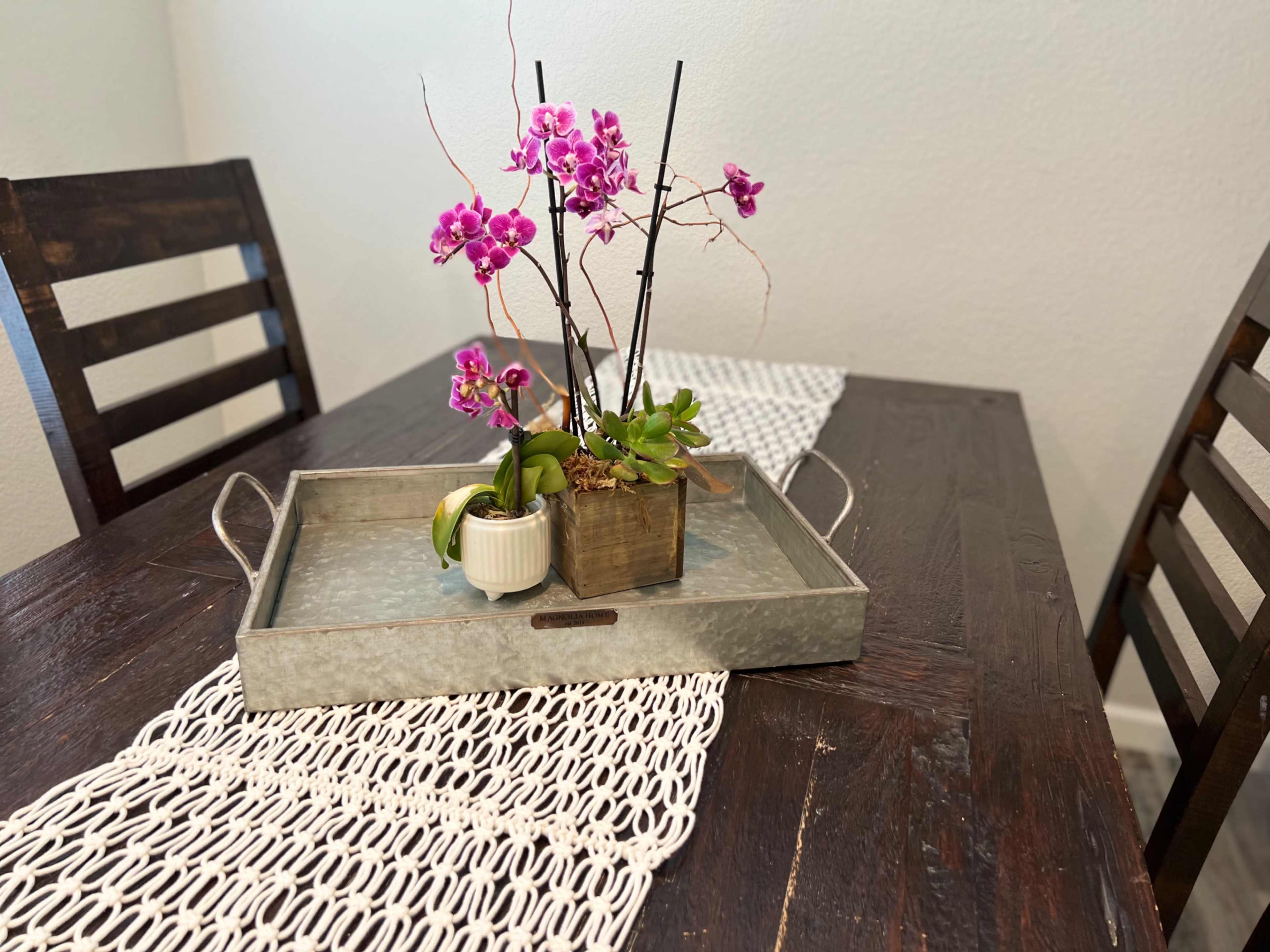A tray with potted orchids and a small succulent is placed on a wooden dining table, flanked by two wooden chairs.