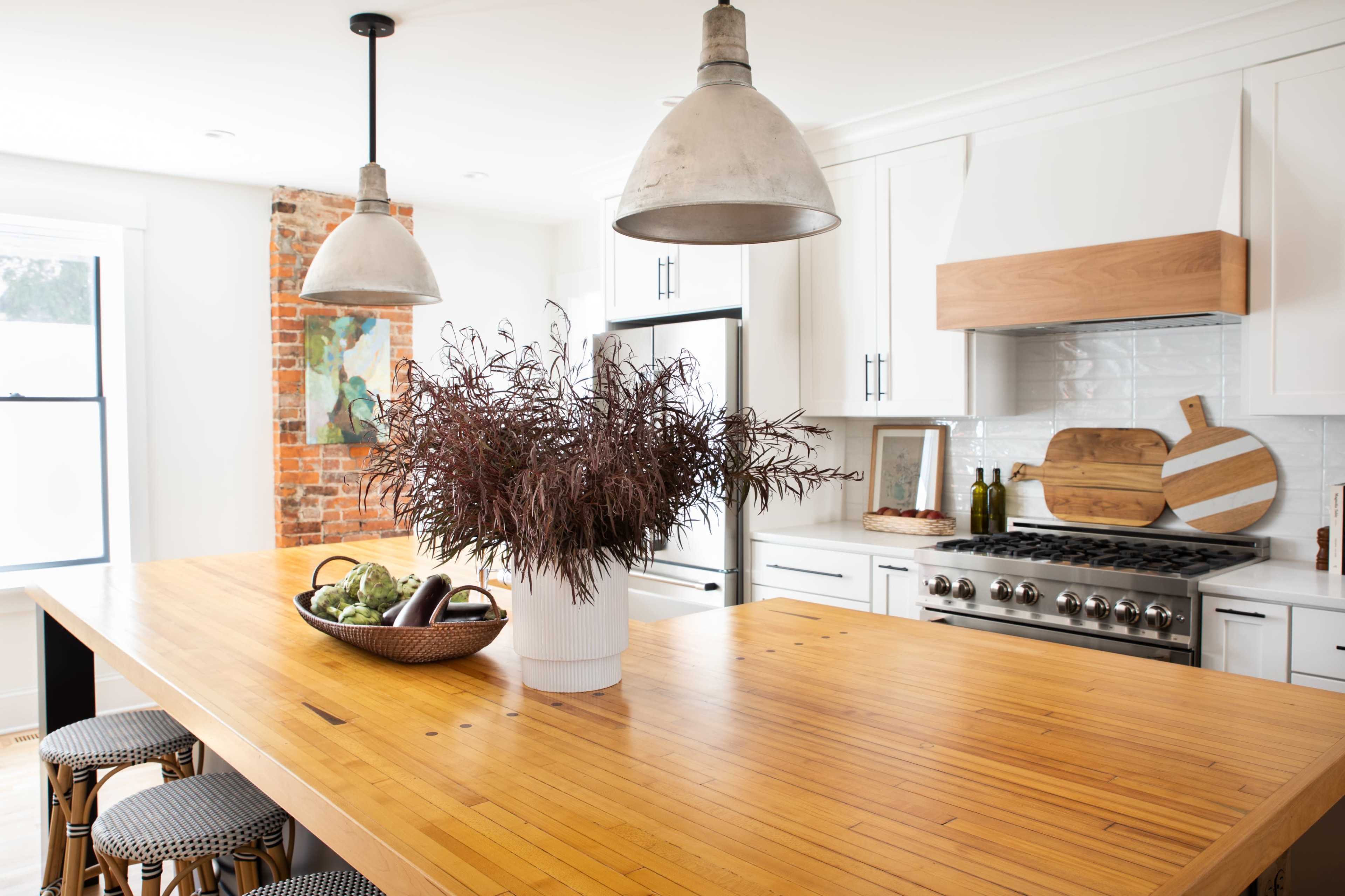 A modern kitchen features a large wooden island with a potted plant at the center, flanked by two pendant lights and surrounded by white cabinets and exposed brick walls.