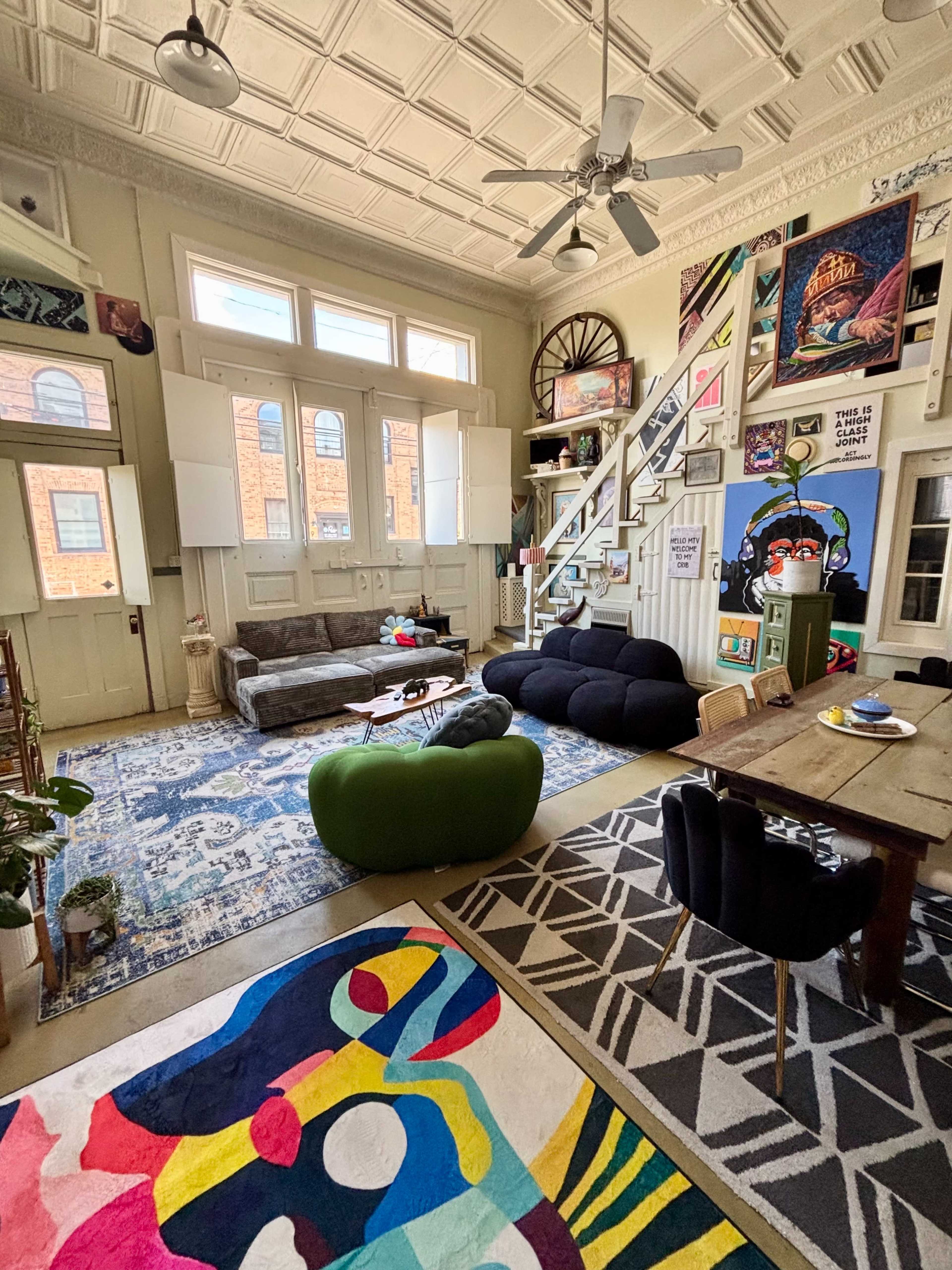 The image shows a spacious living room featuring a mix of colorful art on the walls, a staircase leading to an upper level, and a combination of eclectic furniture, including a green chair and a wooden dining table.