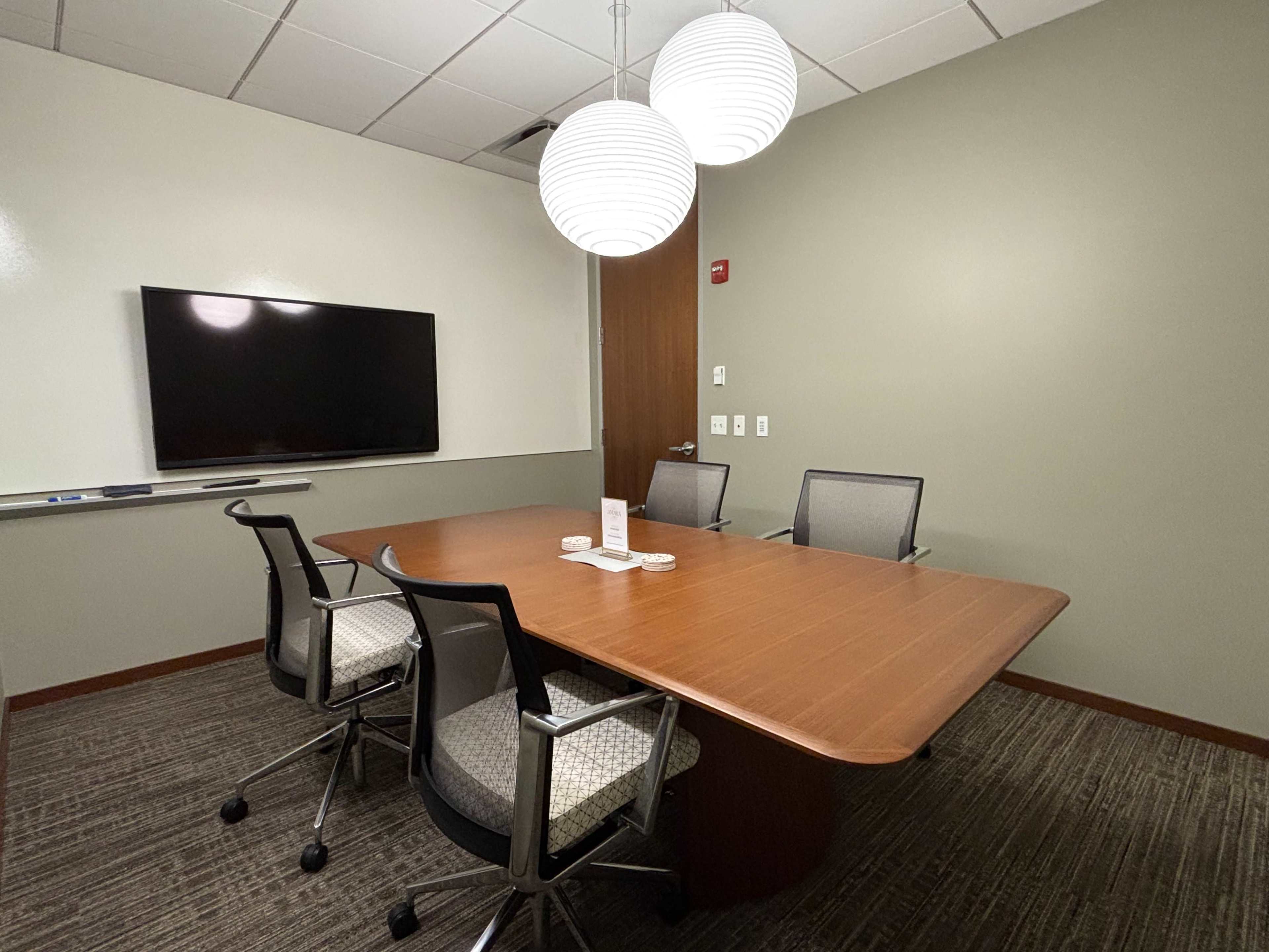 A conference room features a large wooden table surrounded by four chairs, a mounted television on the wall, and two pendant lights overhead.